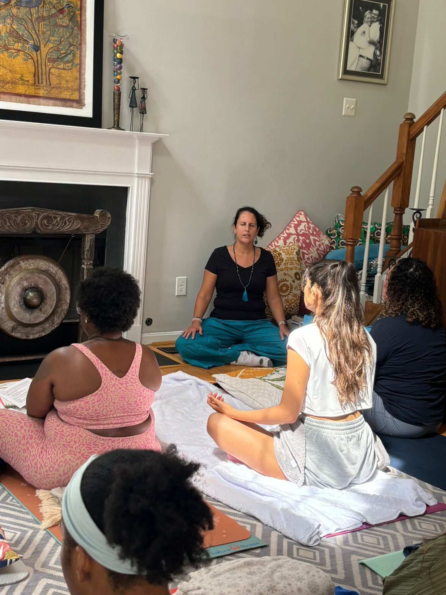 Group of women sitting on mats in a living room, listening to a woman in black speaking, with a fireplace and staircase nearby.