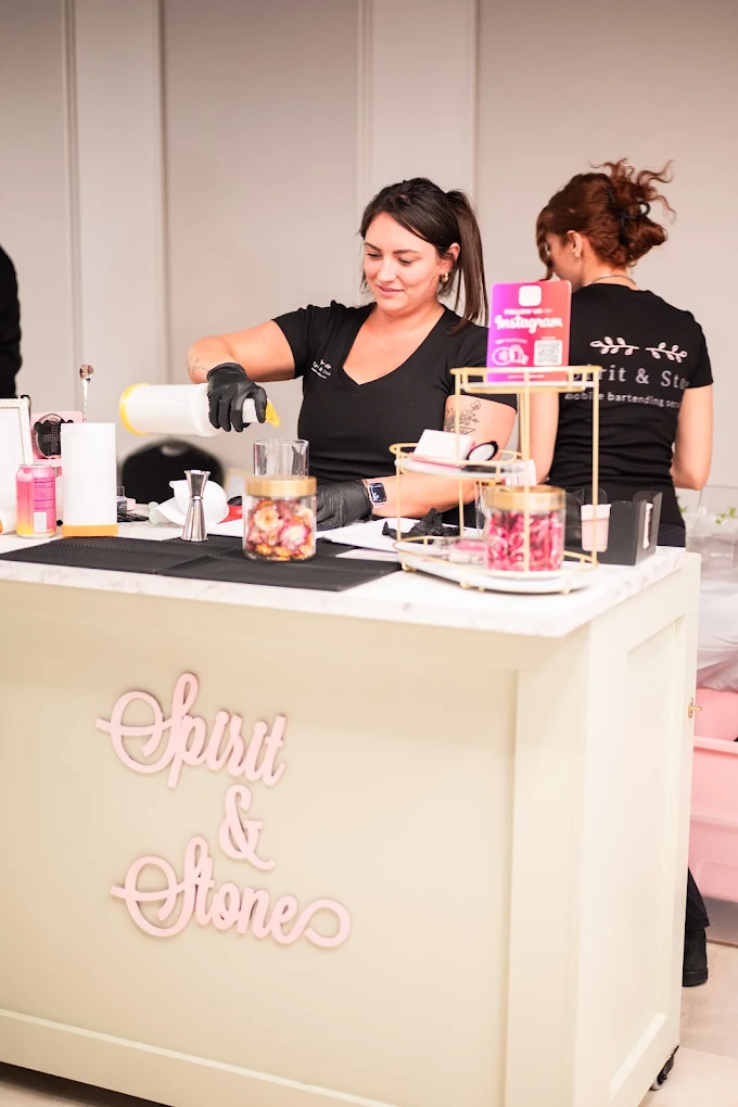 Two women working at a beauty or skincare booth with products and decorations, one smiling and wearing gloves, the other with curly hair.