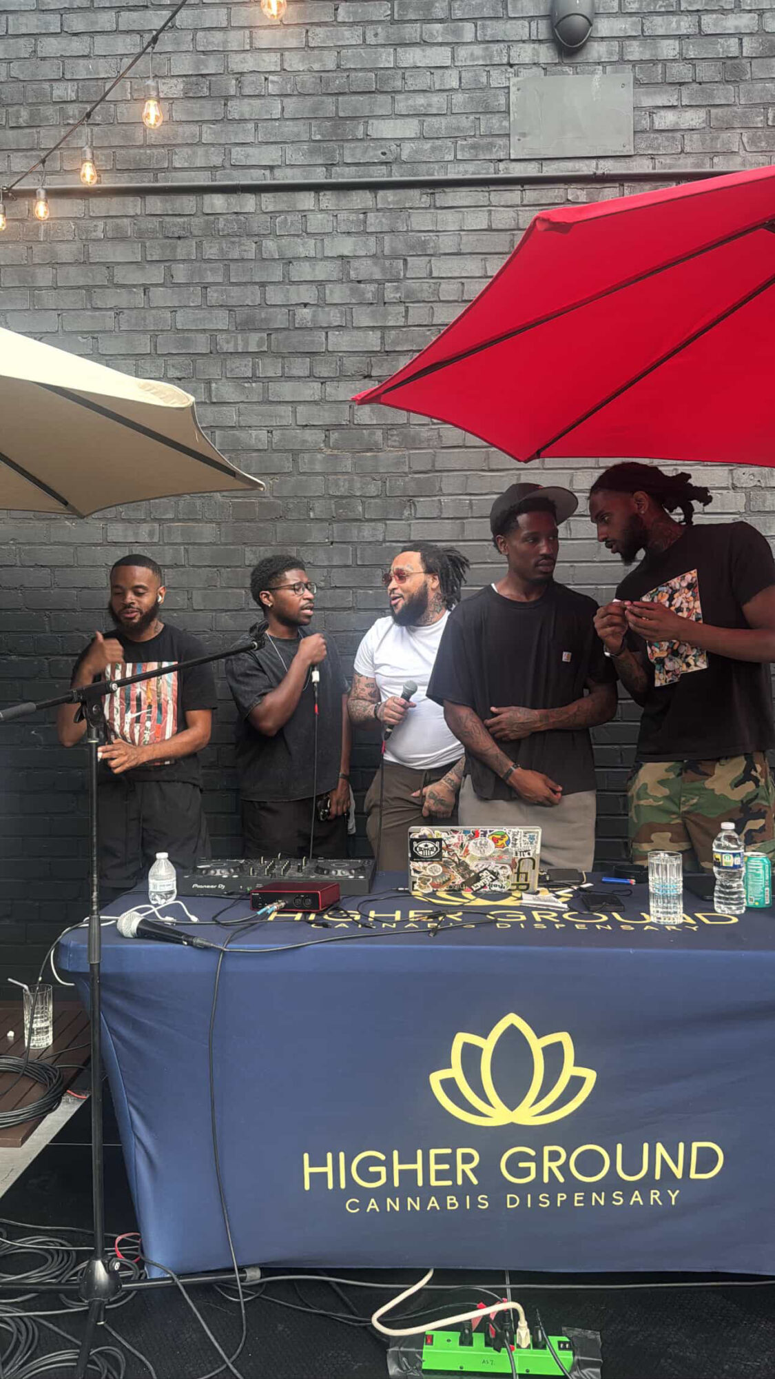 Group of five people standing behind a table with a banner reading 'Higher Ground Cannabis Dispensary' outdoors, with umbrellas overhead.