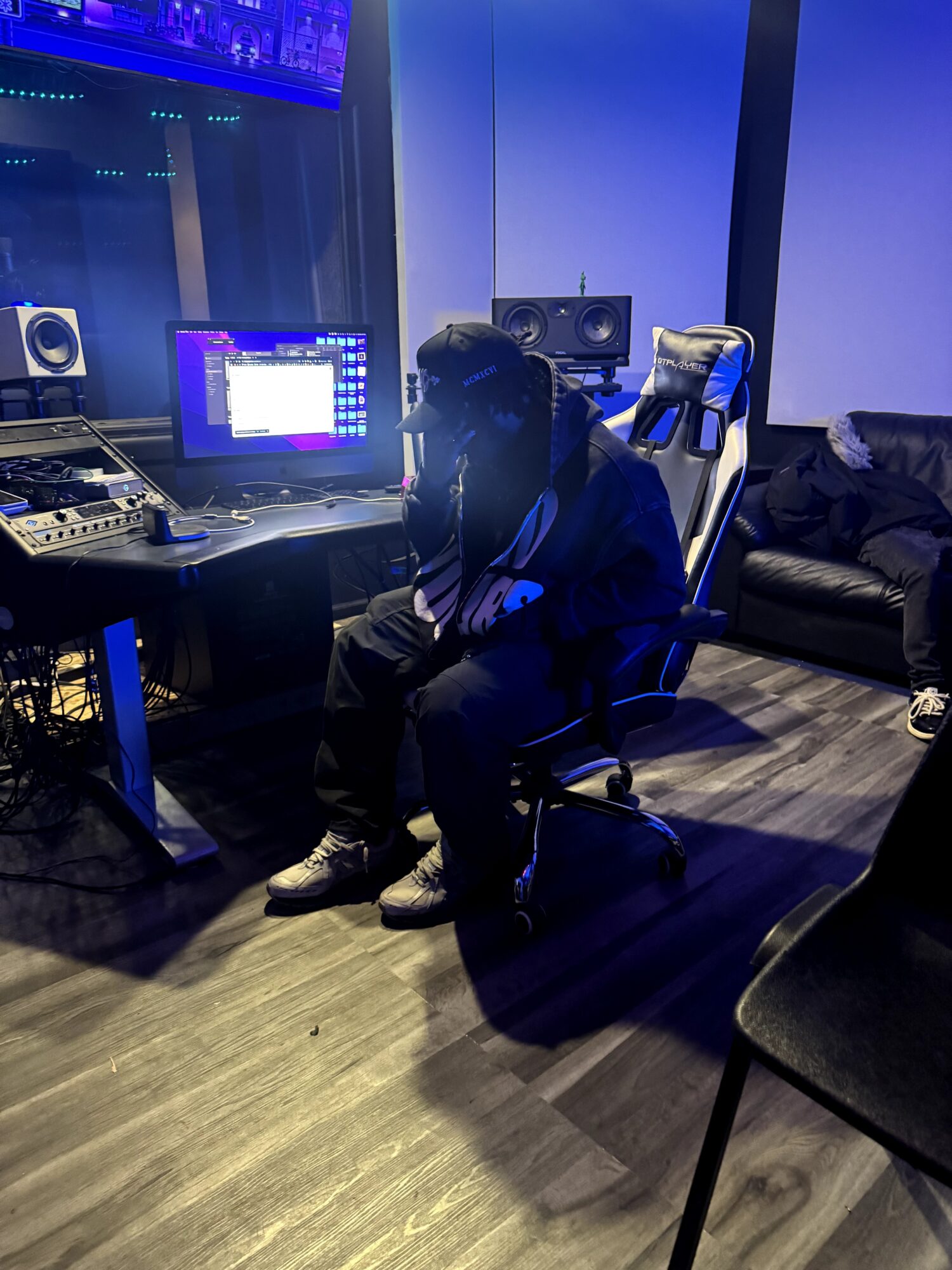 Person sitting in a recording studio with audio equipment and a computer screen, illuminated by blue and purple lighting.