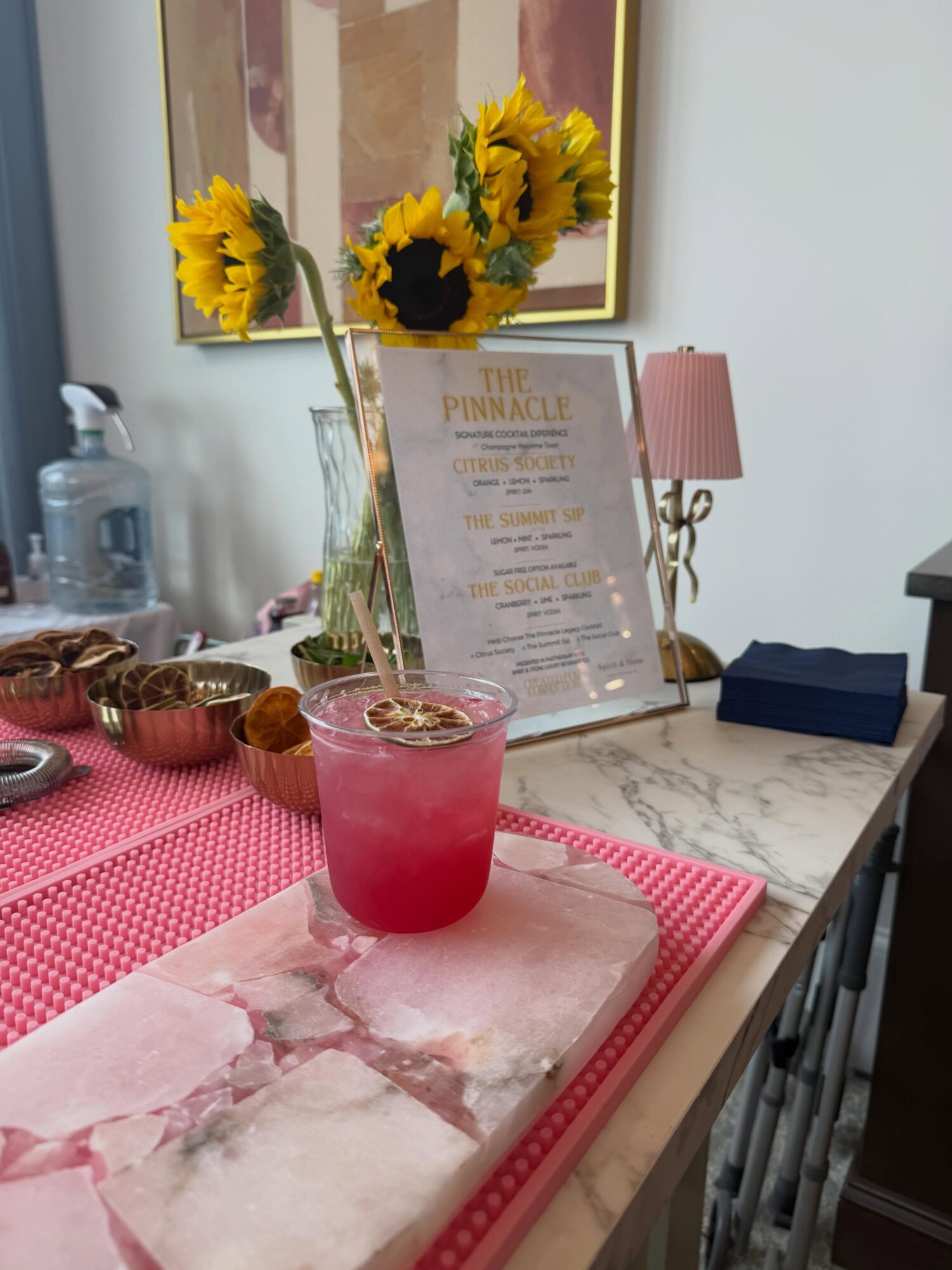 Pink drink in a glass with ice, on a marble cutting board, with sunflowers and a framed menu in the background.