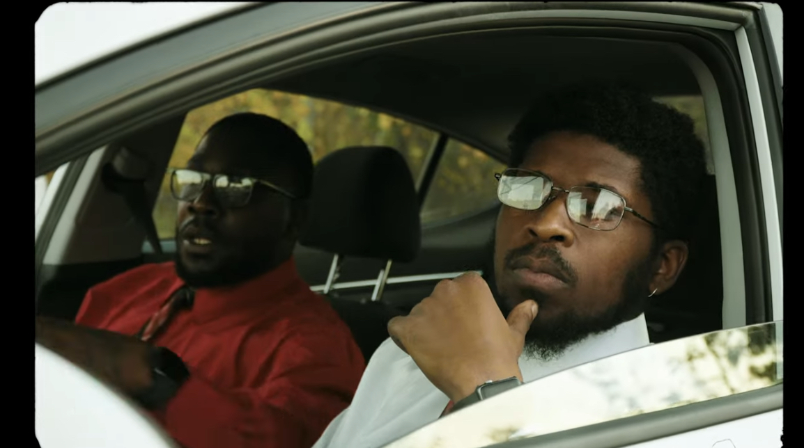 Two men with glasses sitting in a car, one in the driver's seat and the other in the passenger seat.