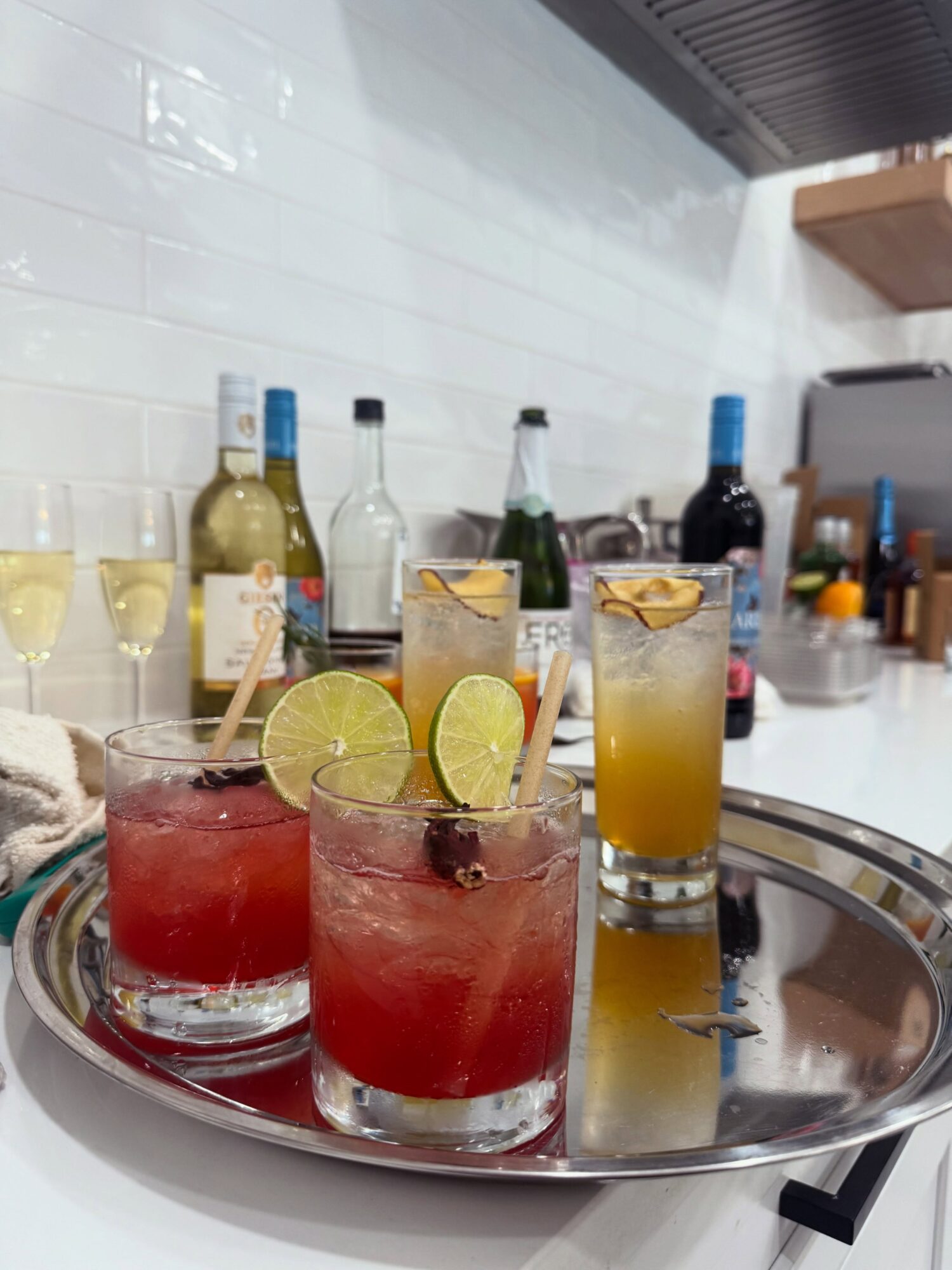 Three red cocktails with lime slices and two yellow drinks with ice on a reflective tray, bar bottles in background.
