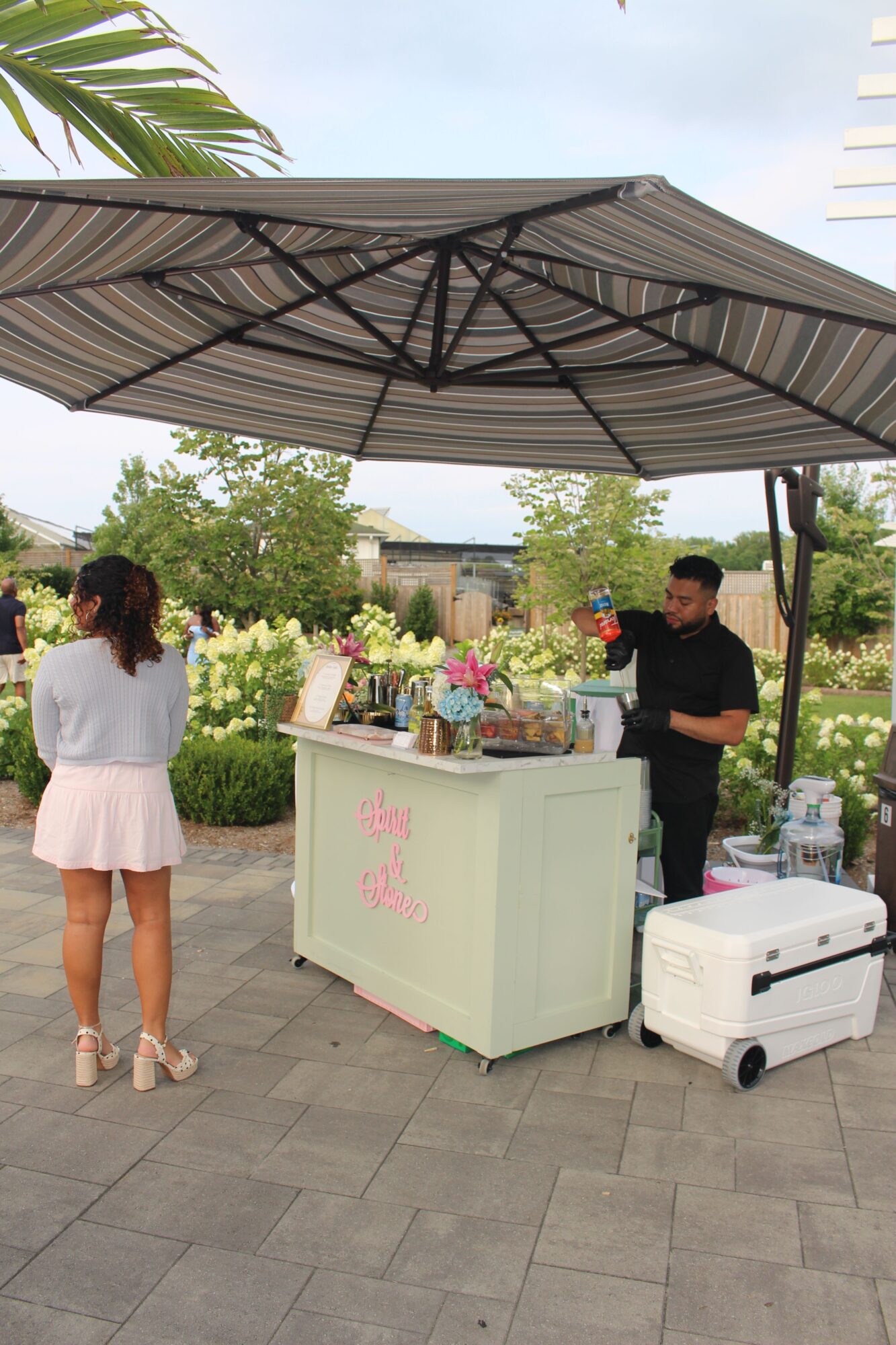 Two women stand outdoors near a table with flowers and a white cooler, under a large umbrella, with trees and sky in background.