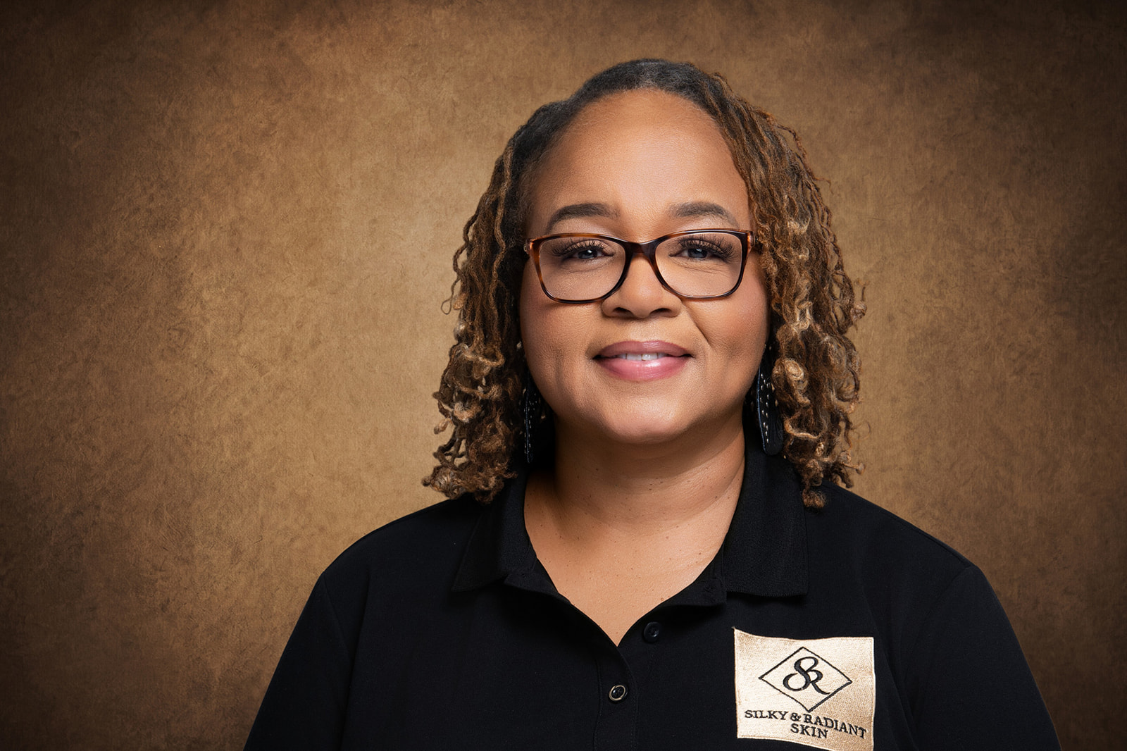 Portrait of a woman with curly hair, glasses, and a black shirt, smiling against a brown background.
