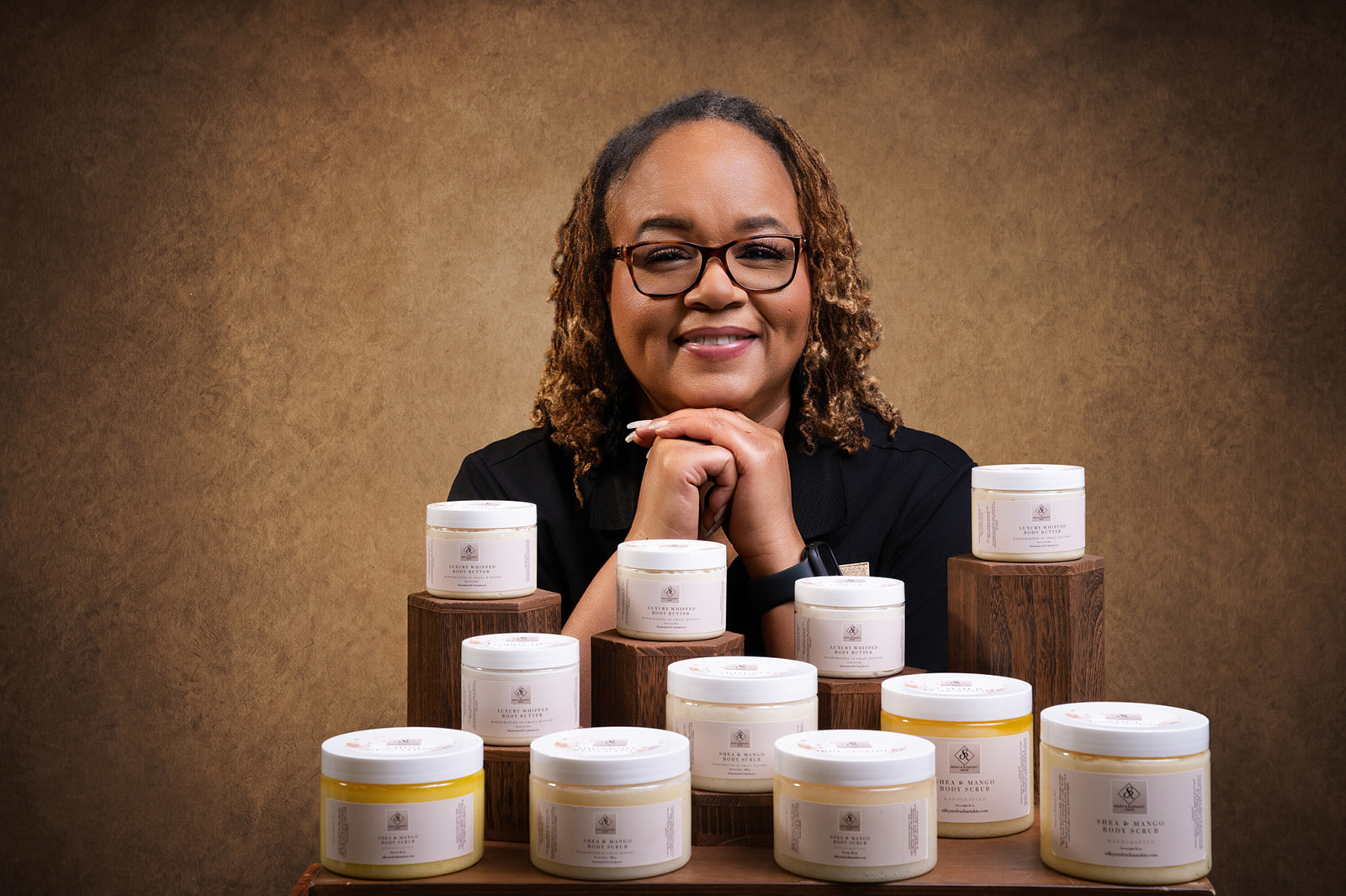 Smiling woman with glasses and curly hair, surrounded by skincare jars on wooden blocks, against a brown background.