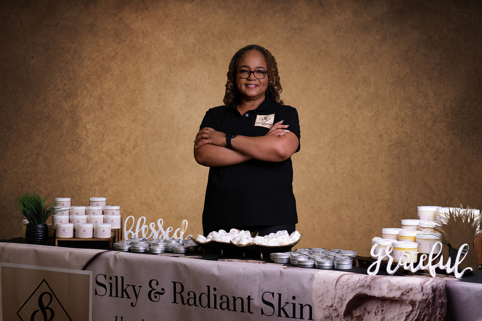 Woman standing behind a table with skincare products, smiling, arms crossed, with