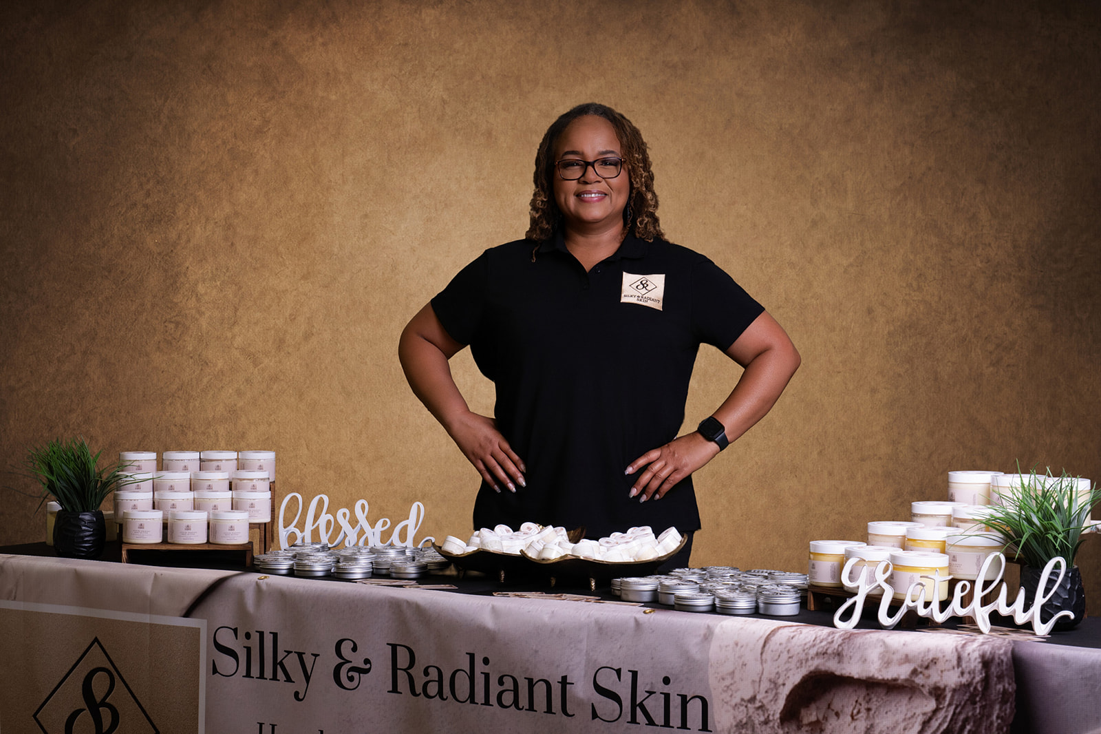Woman standing behind a table with skincare products, smiling, with a gold background, and signs reading 'blessed' and 'grateful'.