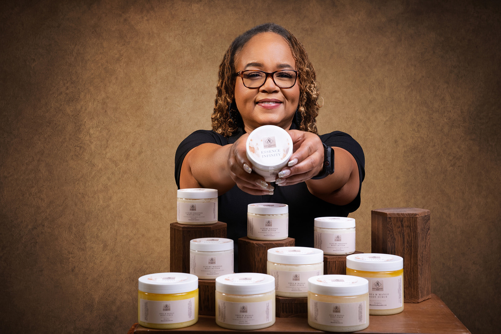 Smiling woman with glasses holding a container, surrounded by similar containers on a wooden table, against a brown background.