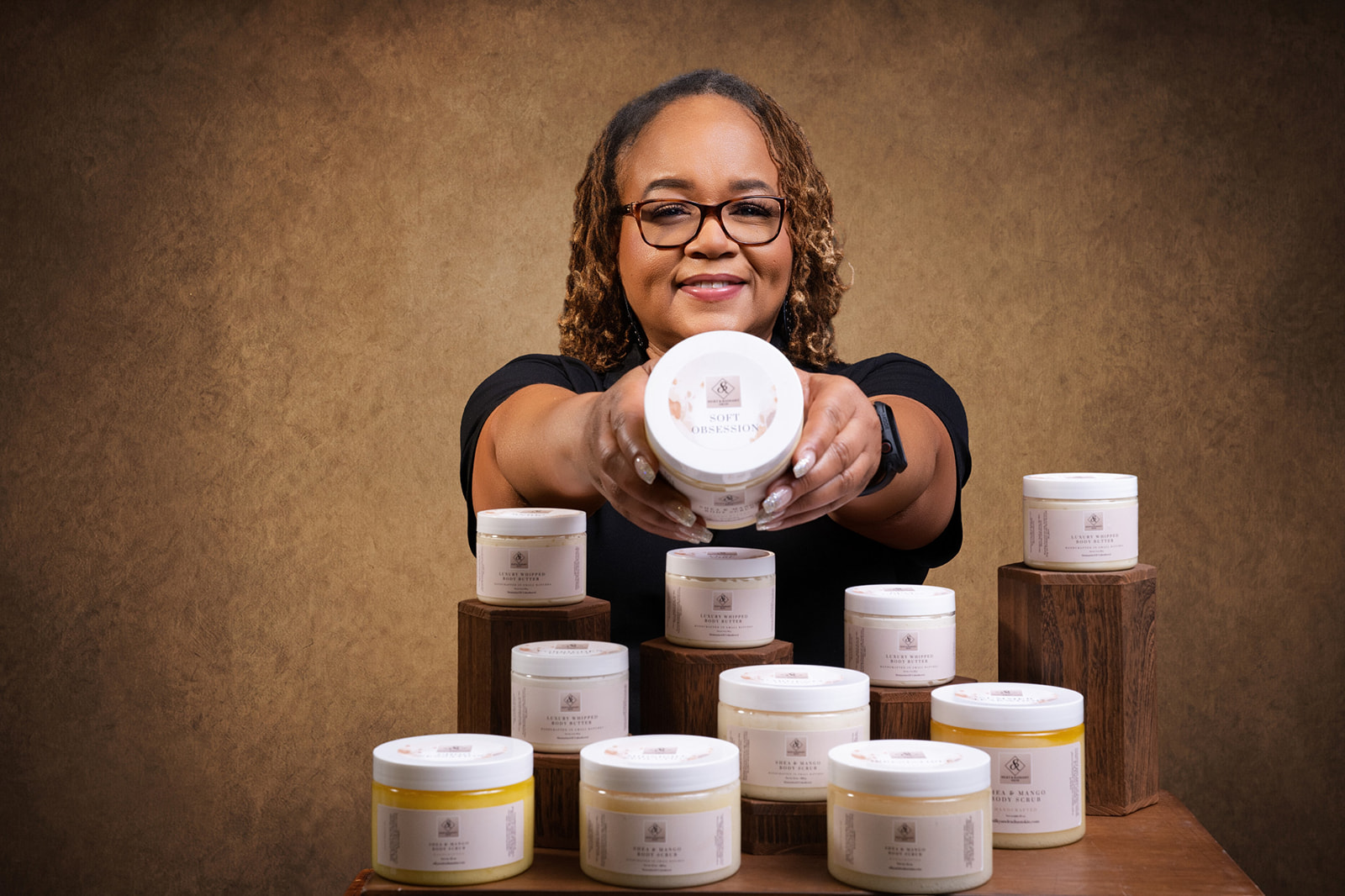Smiling woman with glasses extends hands forward, surrounded by white containers on wooden blocks, against a brown background.
