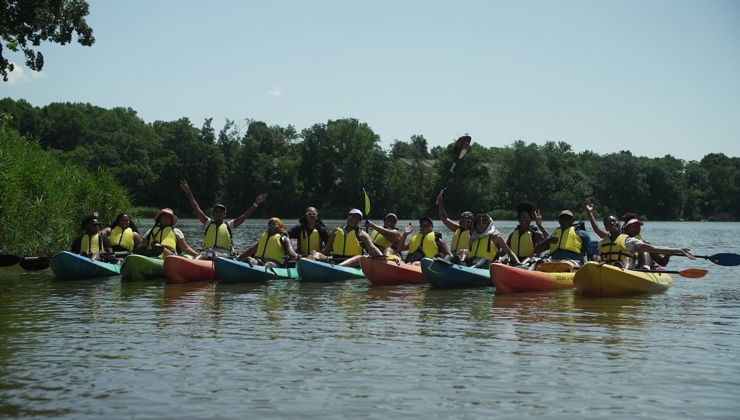 Group of people in kayaks on a calm river, surrounded by trees and greenery, with a clear sky above.