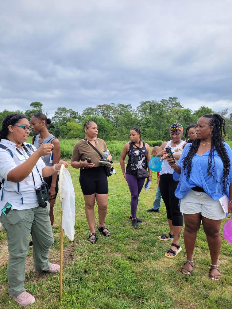 Group of women standing outdoors on grass, engaged in conversation, with trees and cloudy sky in background.