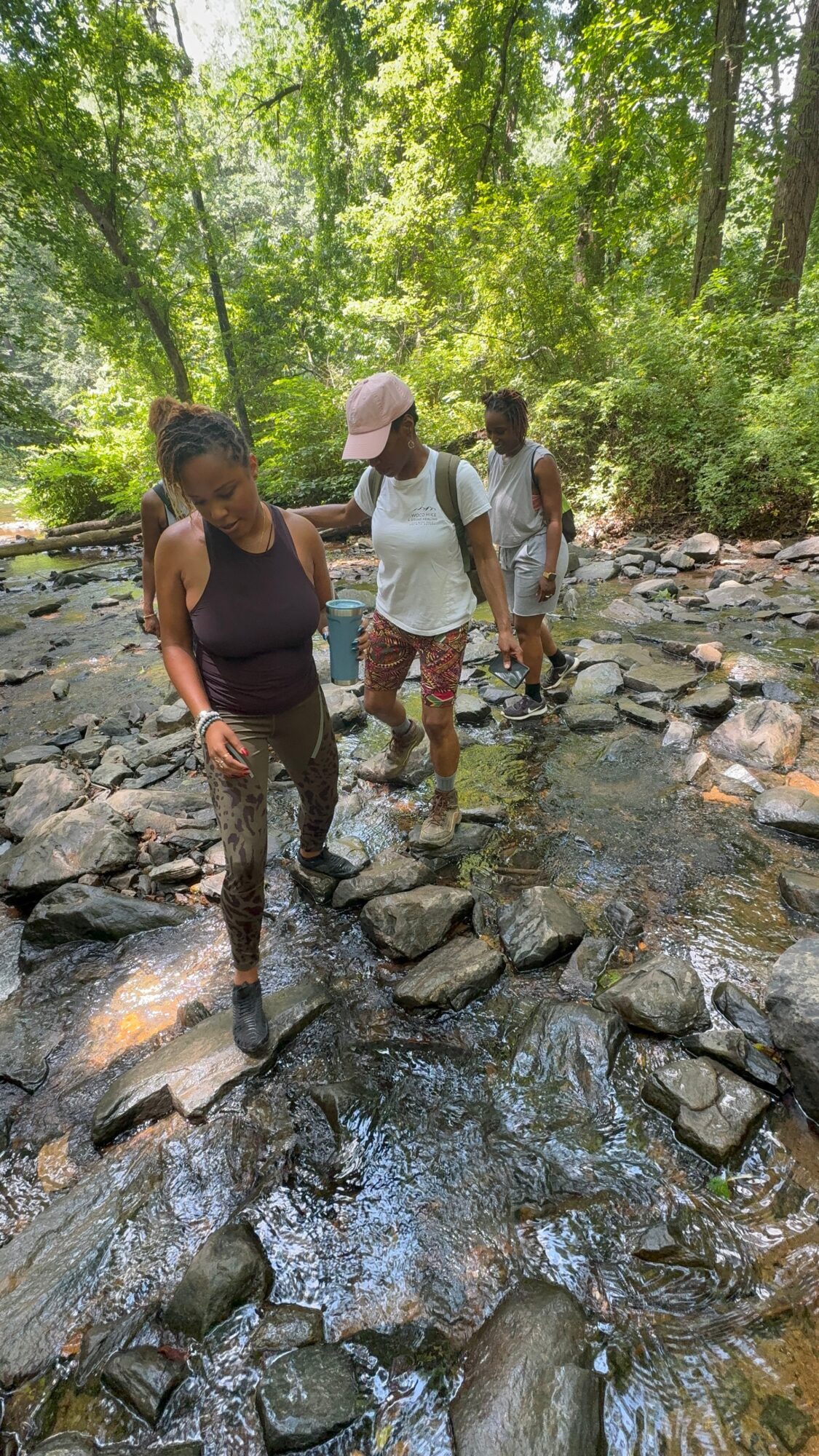 Three people walking across a rocky stream in a forest, surrounded by green trees and sunlight filtering through leaves.