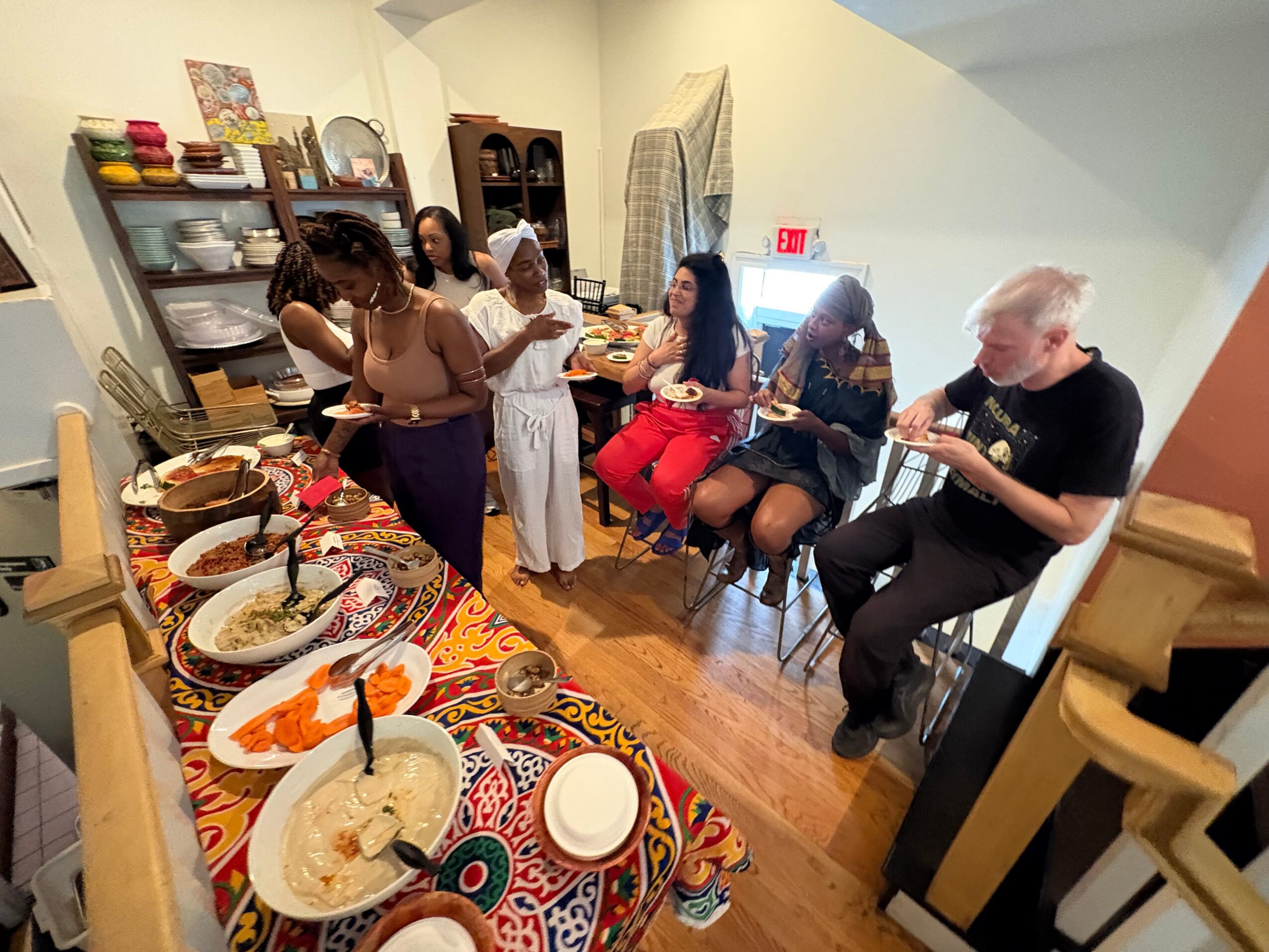 Group of people gathered around a table with food in a room with shelves and a cabinet, some sitting and some standing.