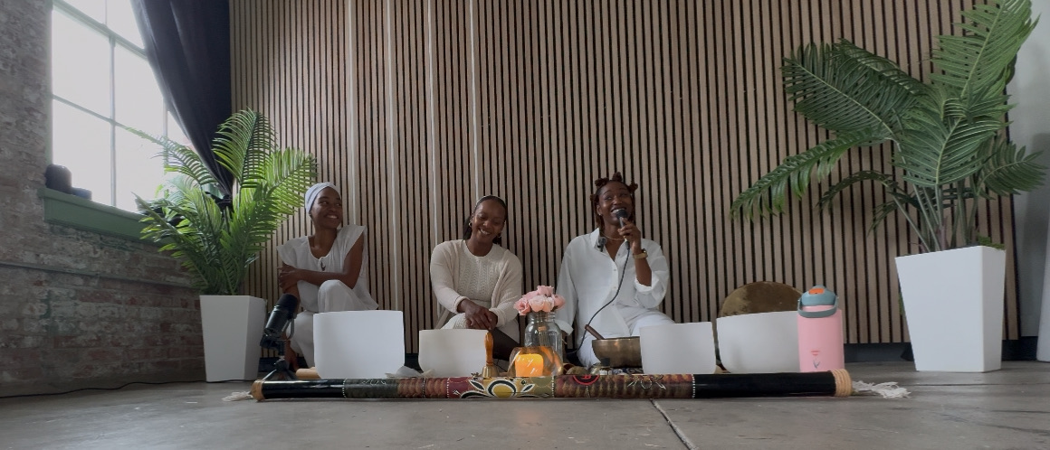 Three people sitting on a white sofa in a modern room with large plants and wooden wall panels, engaging in conversation.