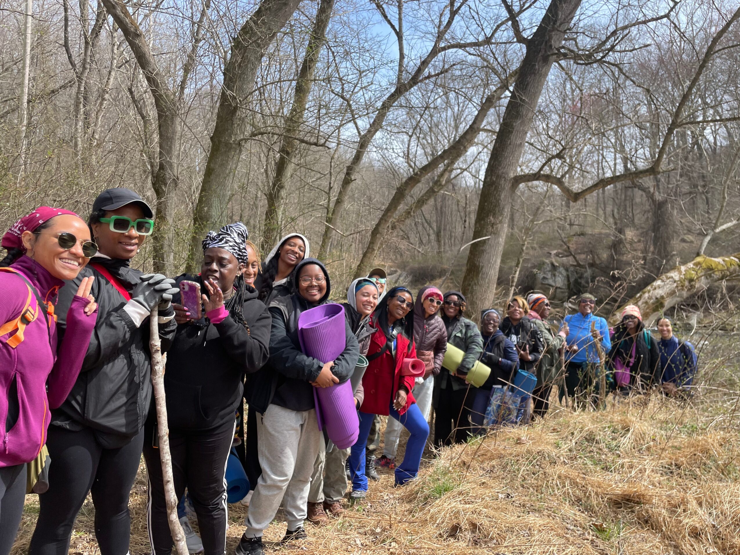Group of people standing in a line outdoors among trees, some holding yoga mats, dressed in outdoor clothing.