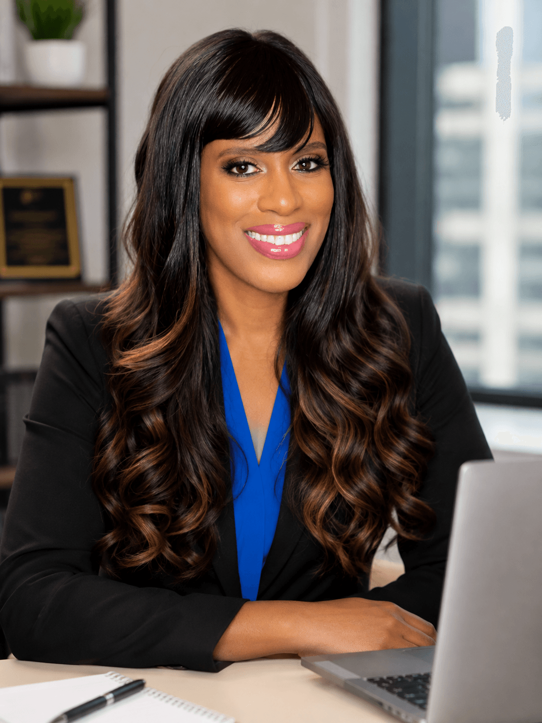Woman with long wavy hair smiling, sitting at desk with laptop in office setting.