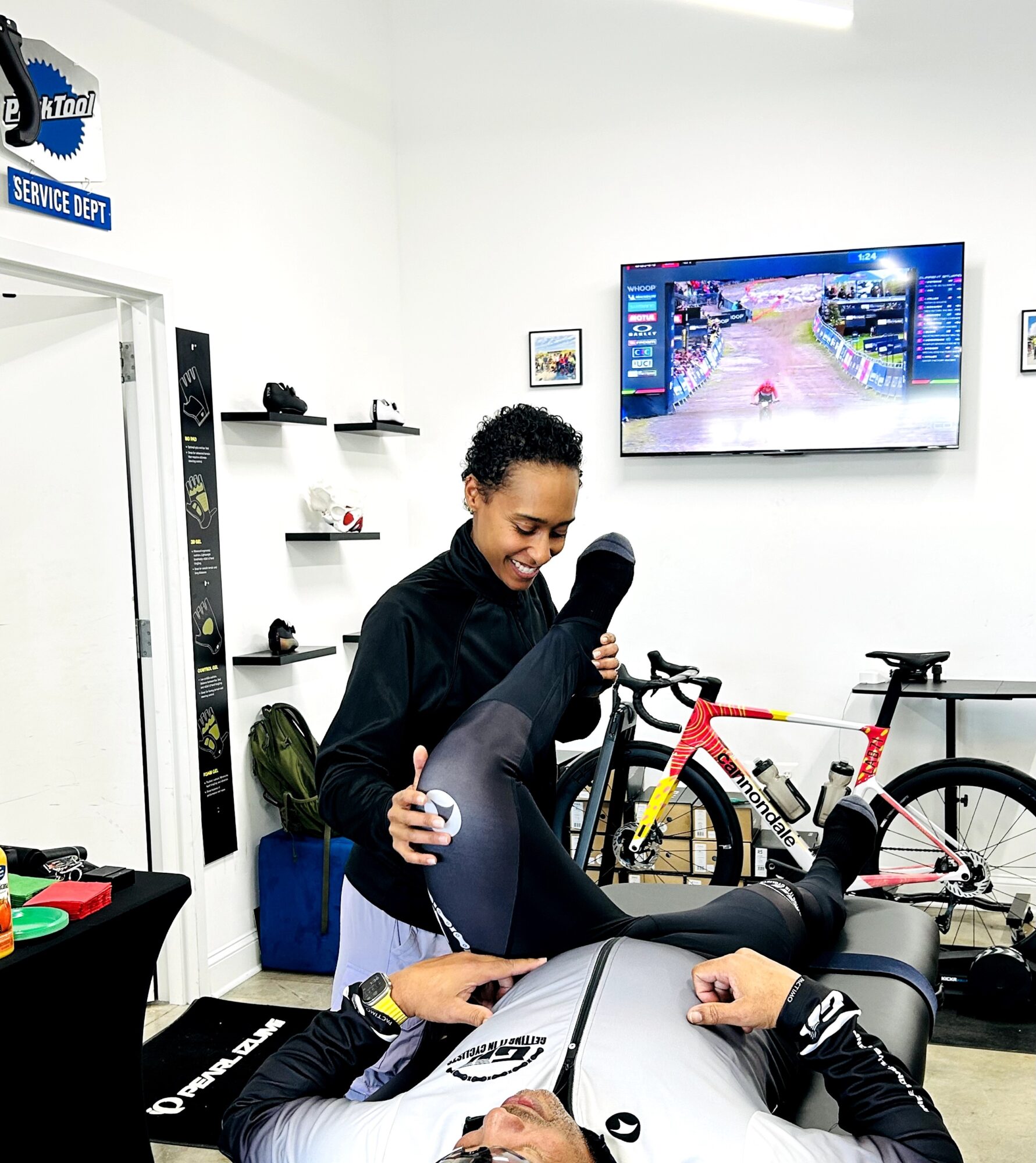 A woman smiling and holding a person's leg in a bike shop with bicycles and a TV screen in the background.