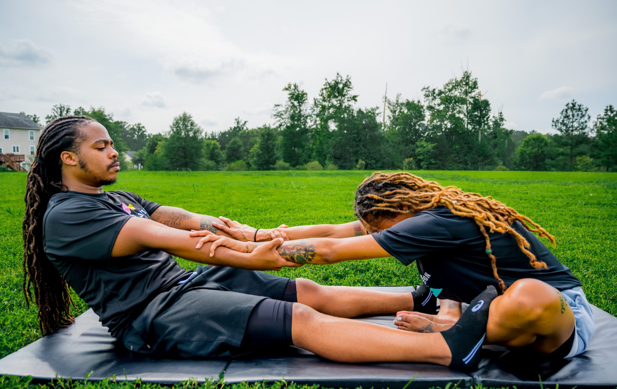 Two people practicing yoga outdoors on a mat, holding hands while sitting on grass with trees in the background.