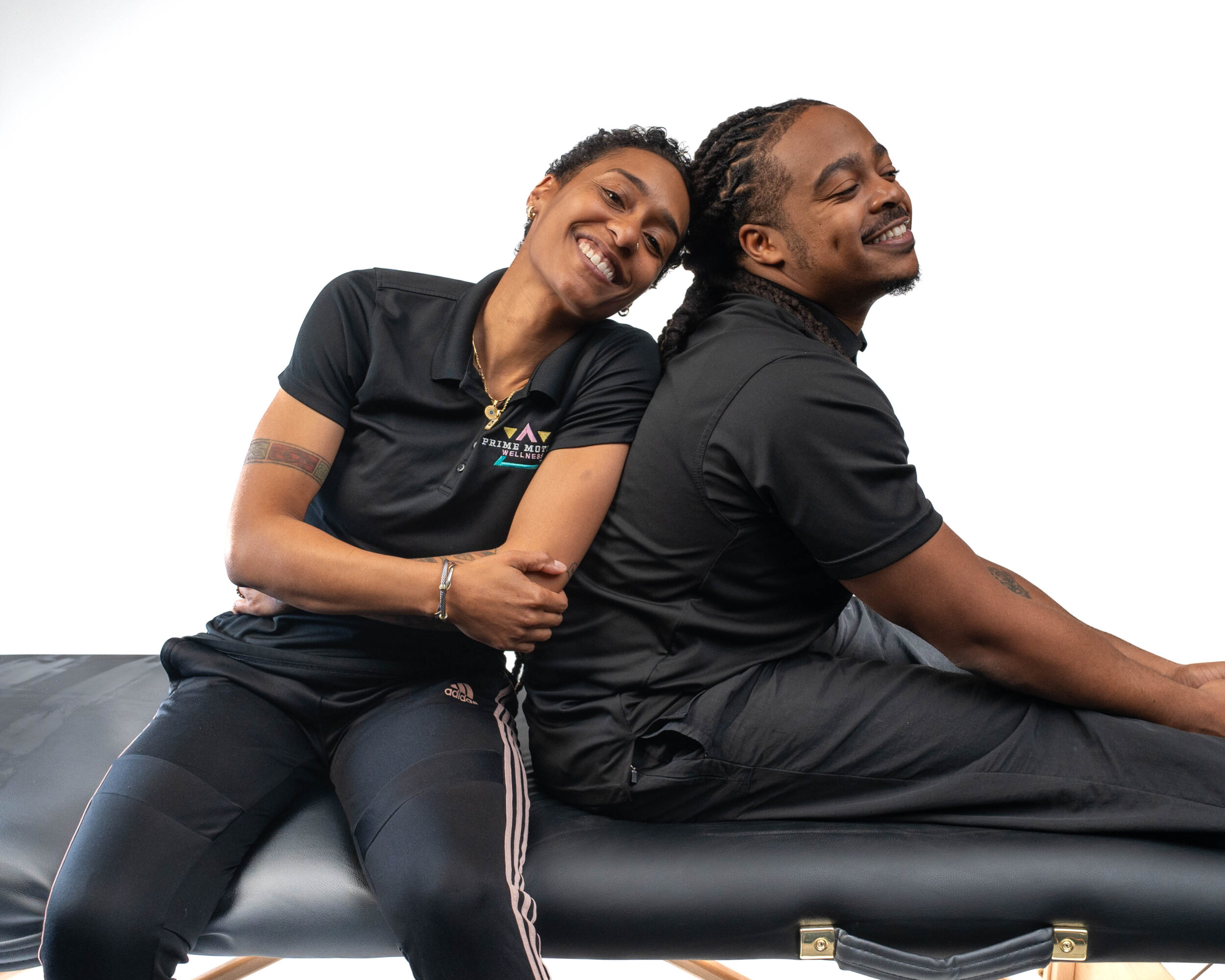 Two people sitting on a black bench, smiling and leaning against each other, with a plain white background.