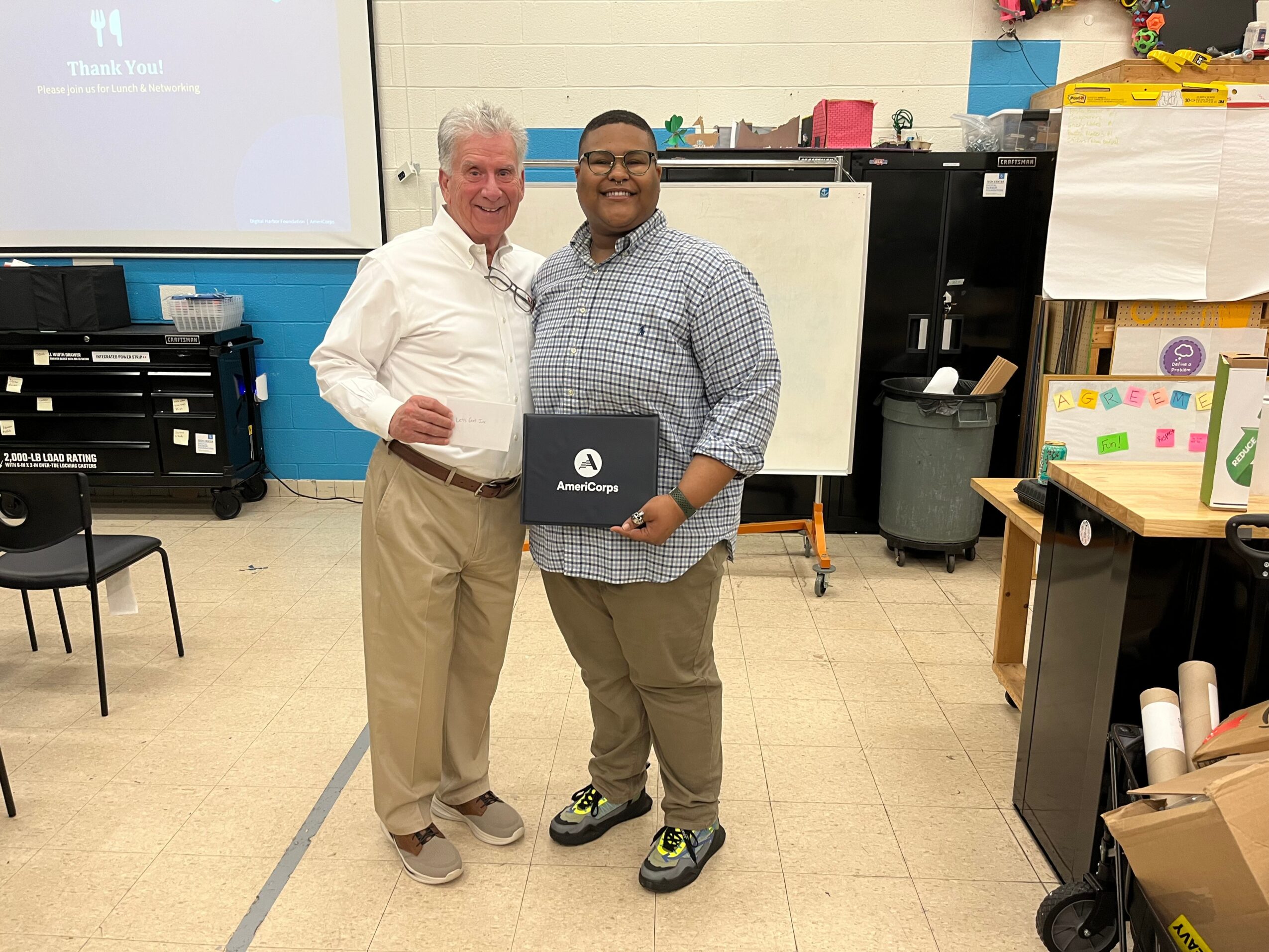 Two men standing in a classroom, smiling, holding awards, with classroom supplies and a whiteboard behind them.
