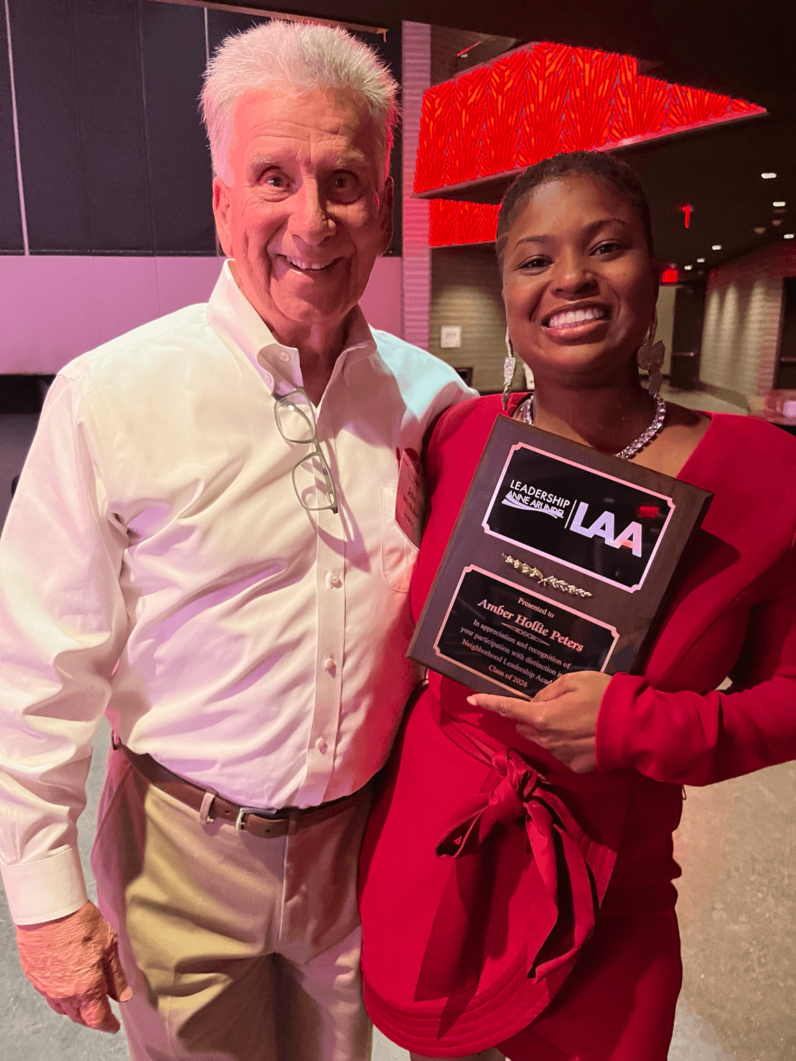 Two people smiling, one holding a plaque, in an indoor setting with red and purple lighting.