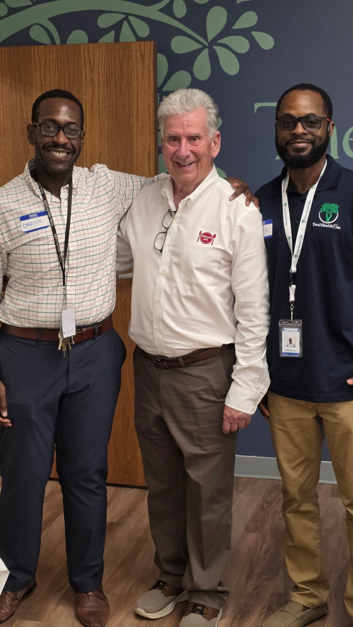 Three men standing together indoors, smiling, with a green leafy wall and wooden door in background.