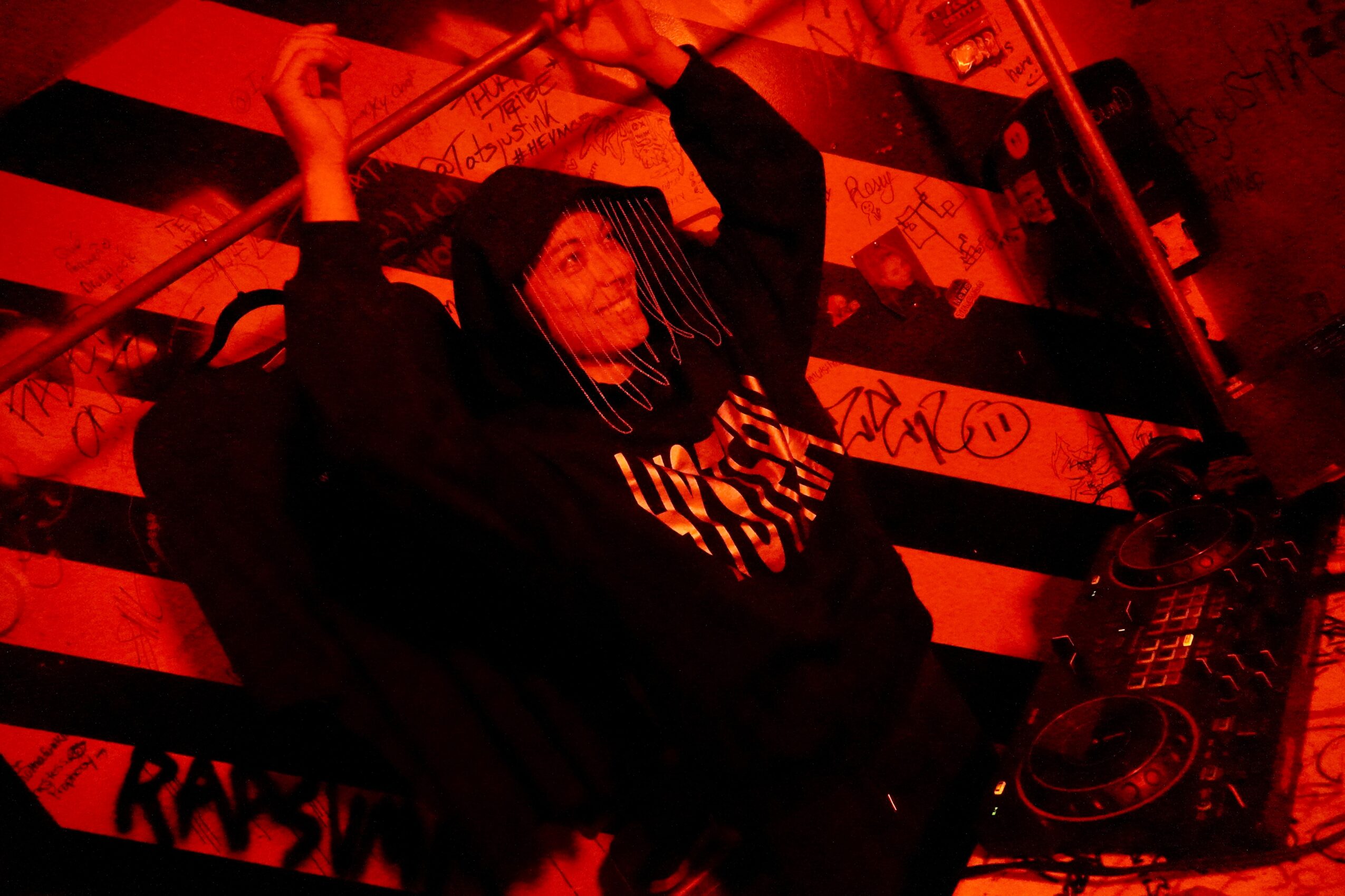Person with raised arms in a dark hoodie, red lighting, graffiti background, and DJ equipment nearby.