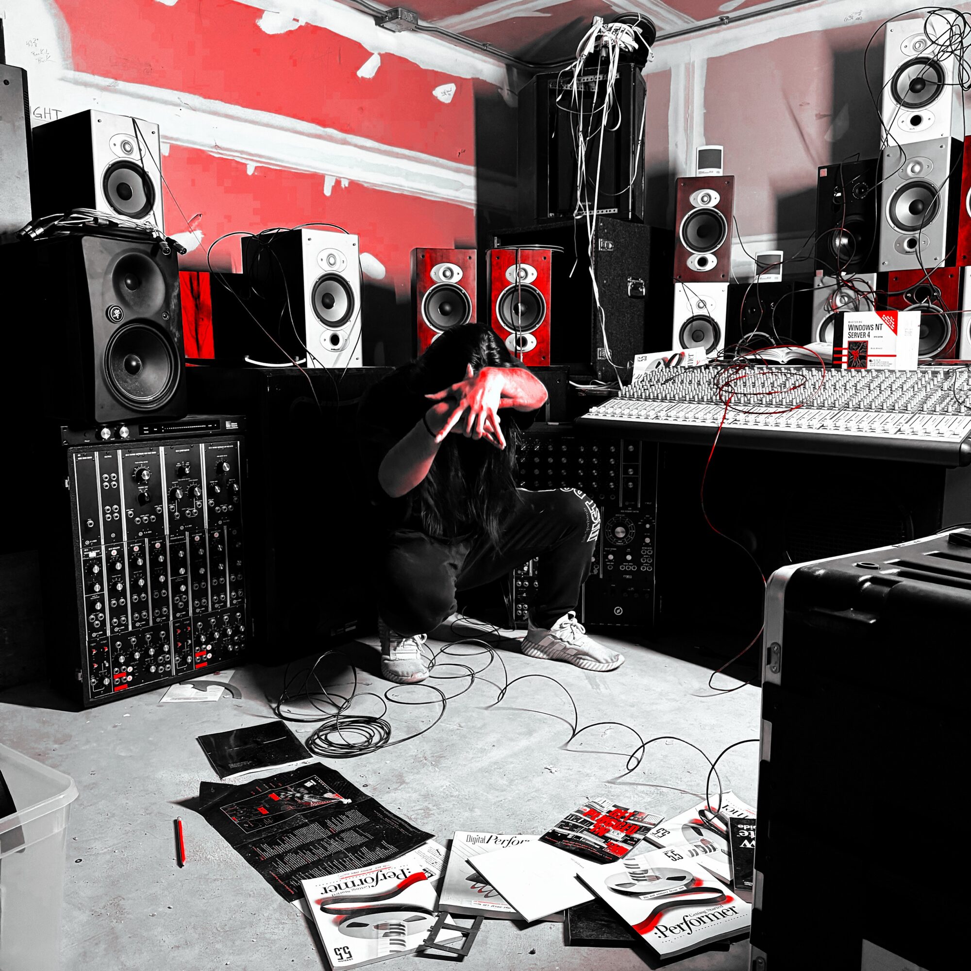 Person crouching in a music studio surrounded by speakers and audio equipment, with papers and cables on the floor.
