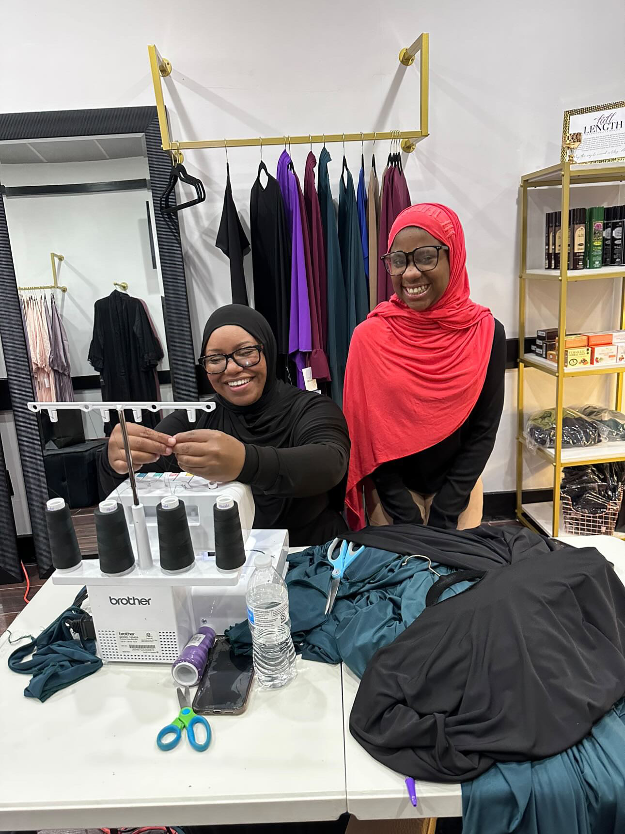 Two women in hijabs smiling in a clothing store, with sewing supplies and clothes on a table.