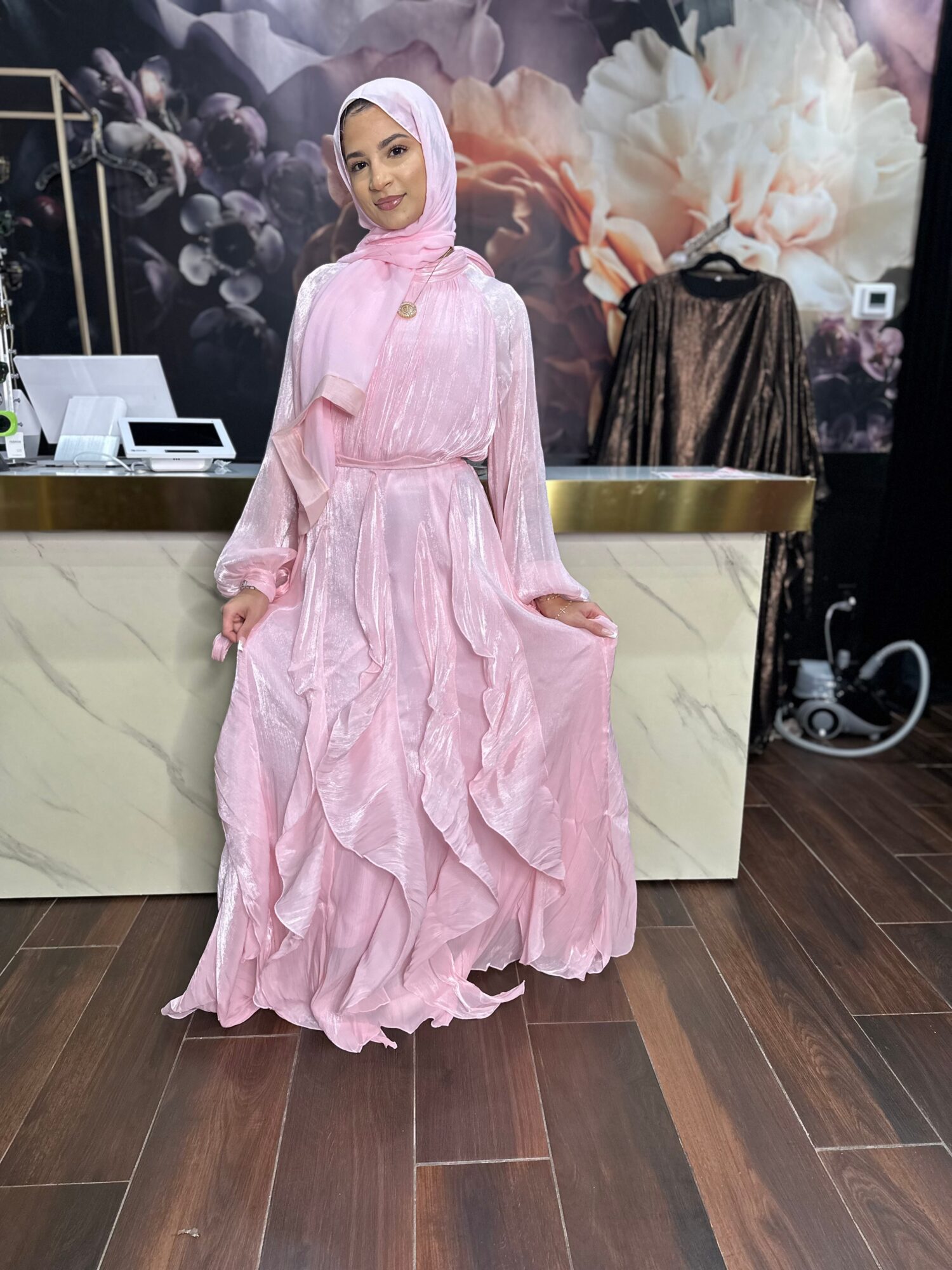 Woman in pink dress and hijab standing indoors with floral artwork background, marble counter, and wooden floor.
