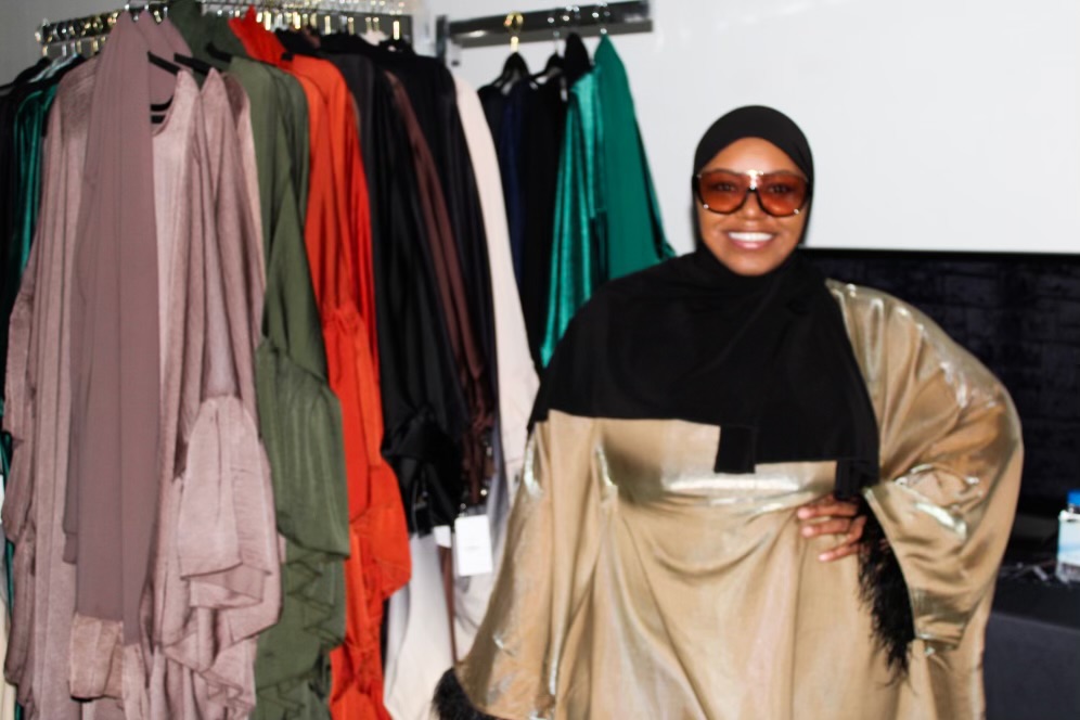 Person wearing sunglasses and a hijab, smiling, standing in front of clothing racks with colorful garments.