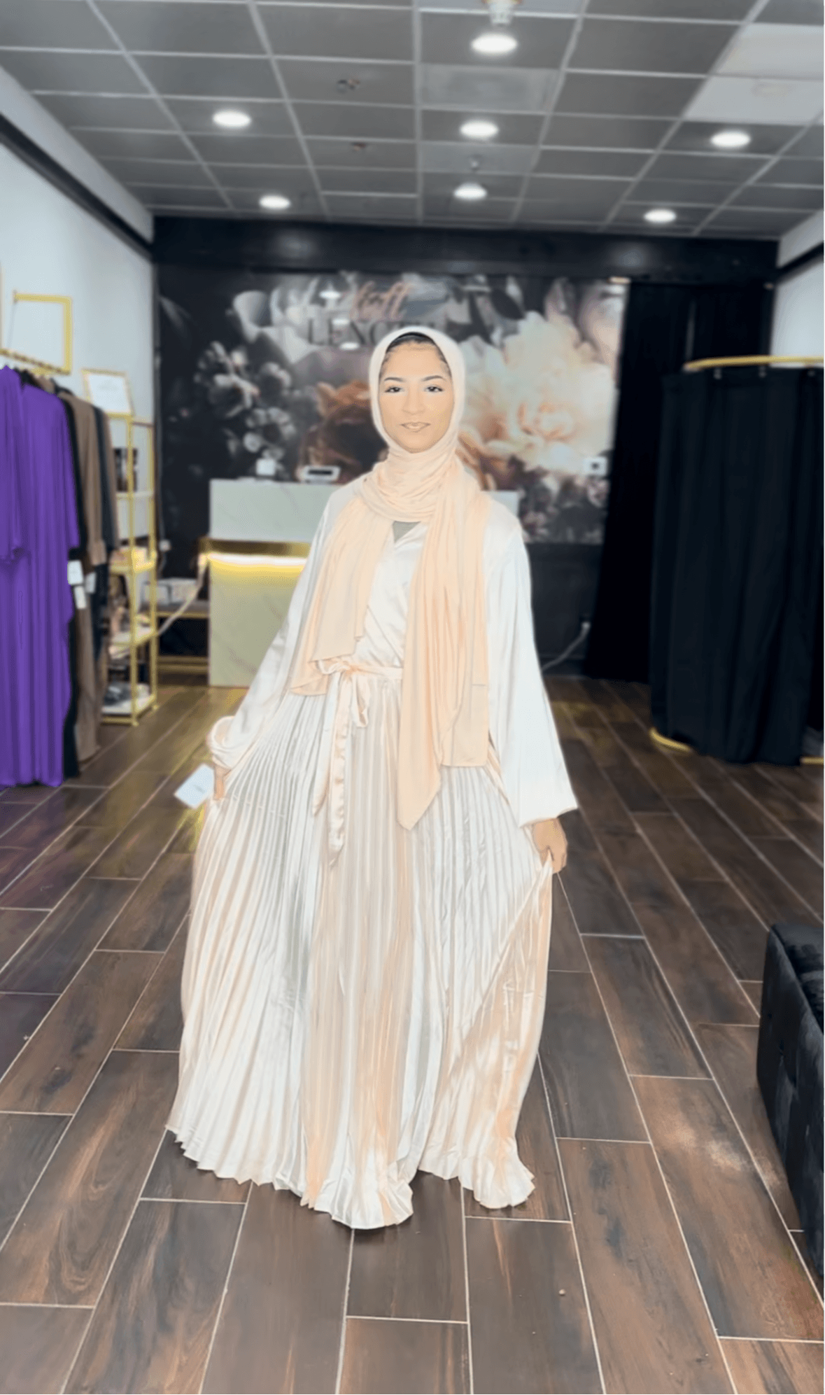 Woman in a long, striped dress and headscarf standing indoors with clothing racks and a dark backdrop behind her.