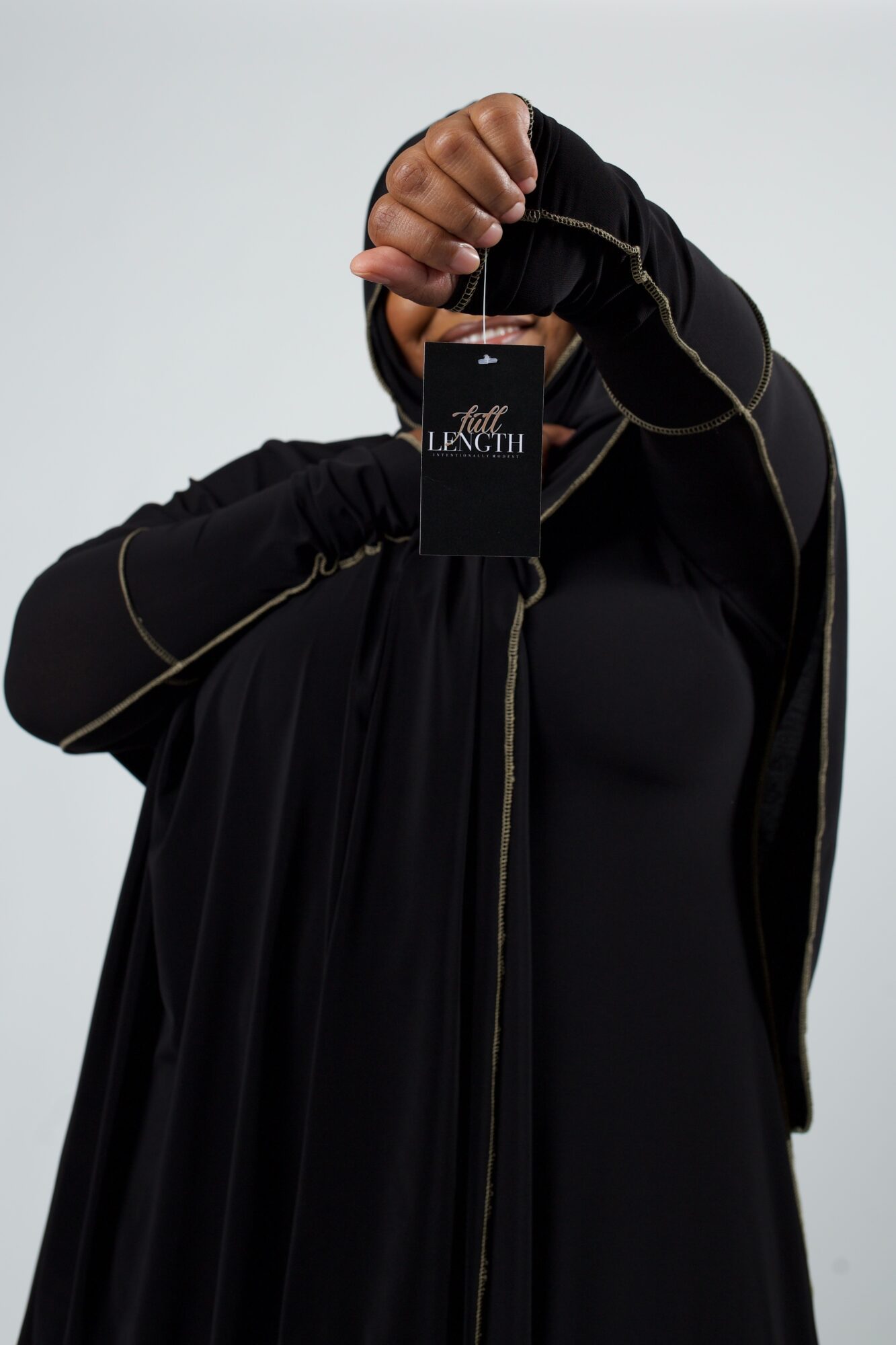 Person holding a black tag with gold text, wearing a black jacket with gold zipper details, against a plain background.