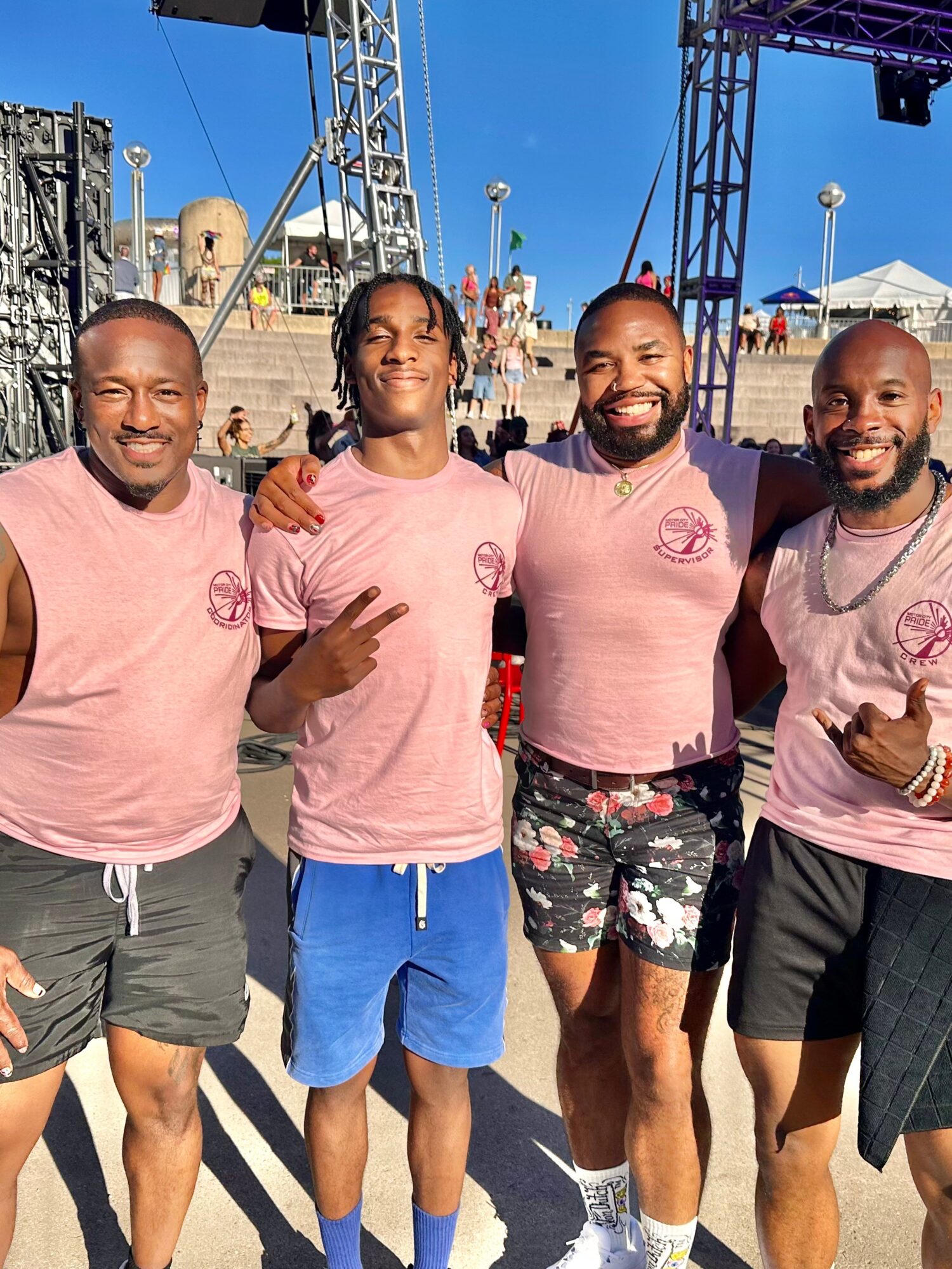 Four men standing outdoors, smiling, wearing matching pink shirts, with a stage and structures in the background.