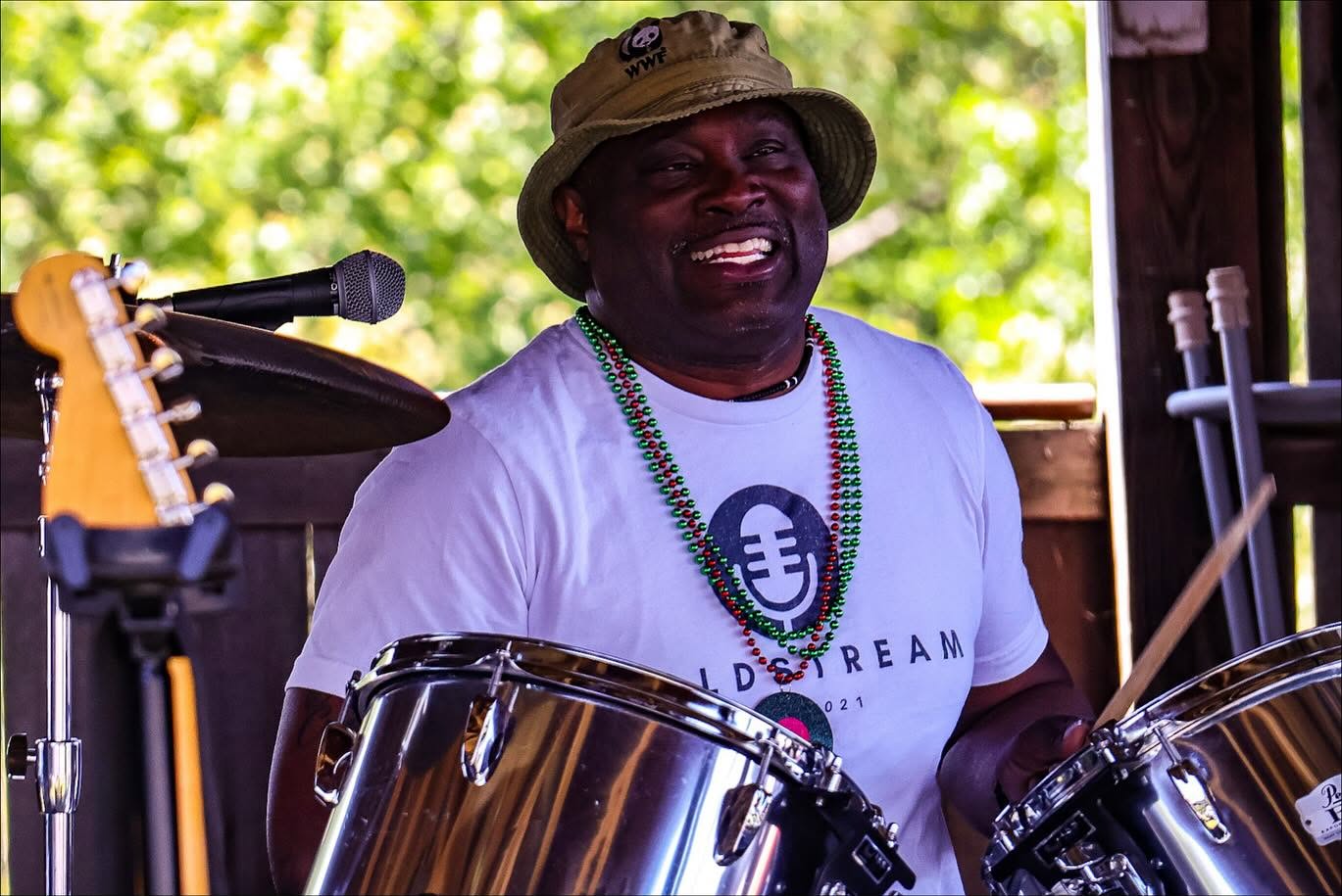 Person playing drums, wearing a hat and a white T-shirt, smiling, with a microphone and musical equipment, outdoors.