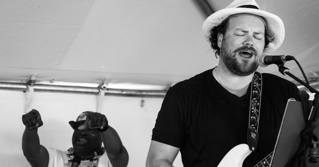 Musician with a hat singing into a microphone, with a person in the background raising fists.