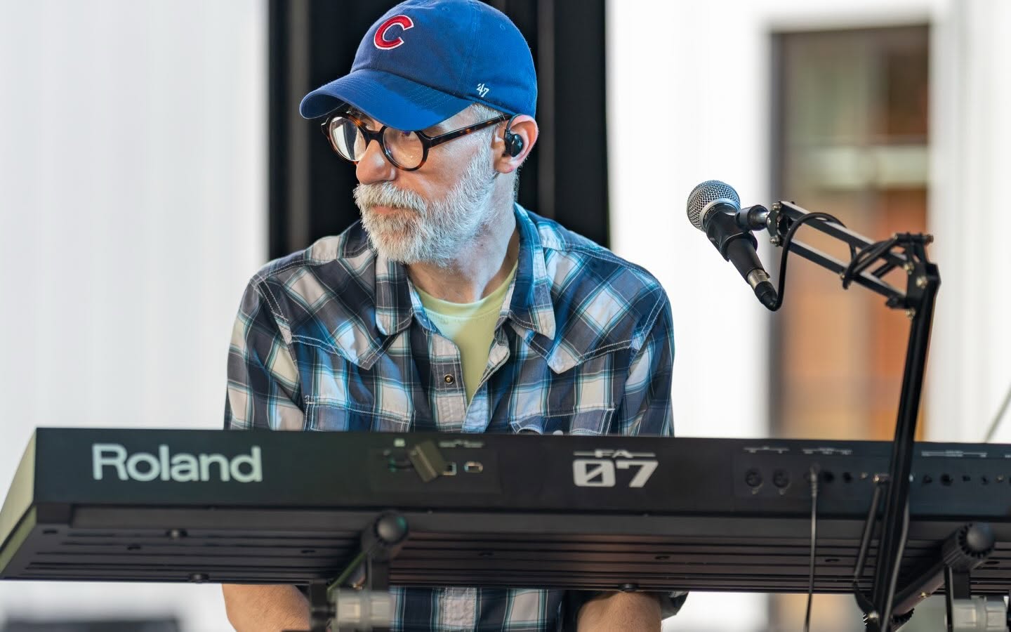 Older man with glasses and a beard wearing a blue cap and plaid shirt playing a keyboard, microphone nearby.