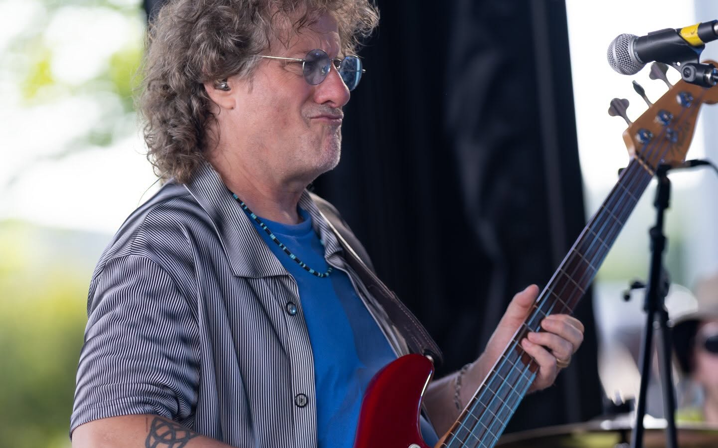 Man with curly hair and glasses playing an electric guitar outdoors, wearing a striped shirt and blue t-shirt.