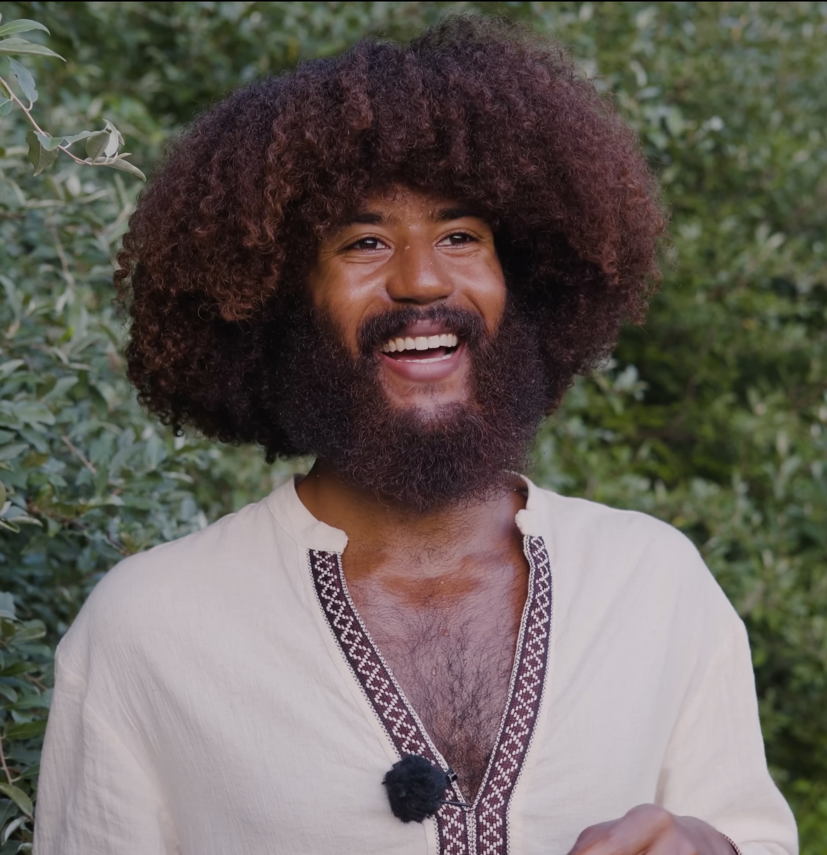 Smiling man with curly hair and beard outdoors, wearing a light-colored shirt with embroidery, surrounded by greenery.