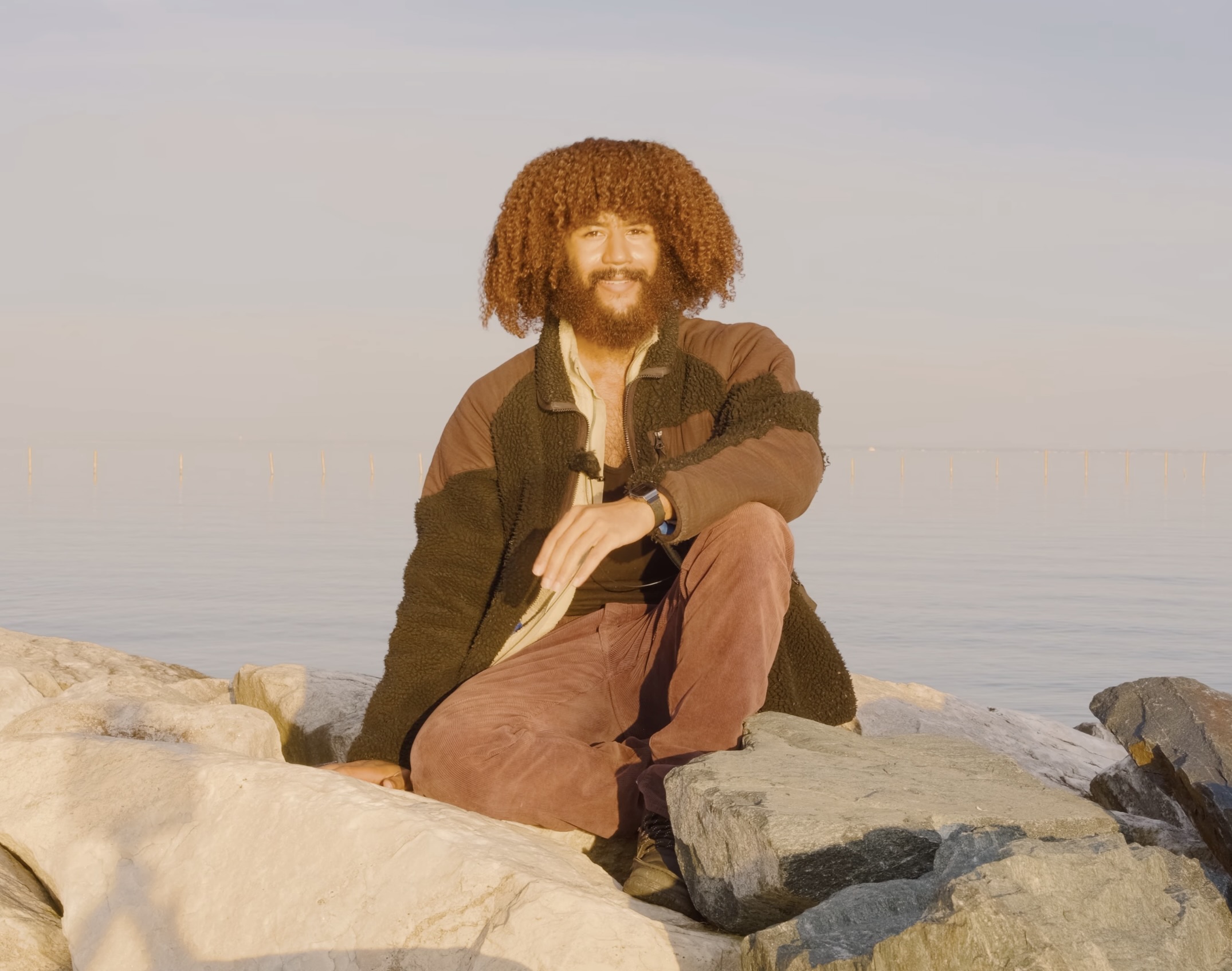 Person with curly hair and beard sitting on rocks near water, smiling, during daytime.