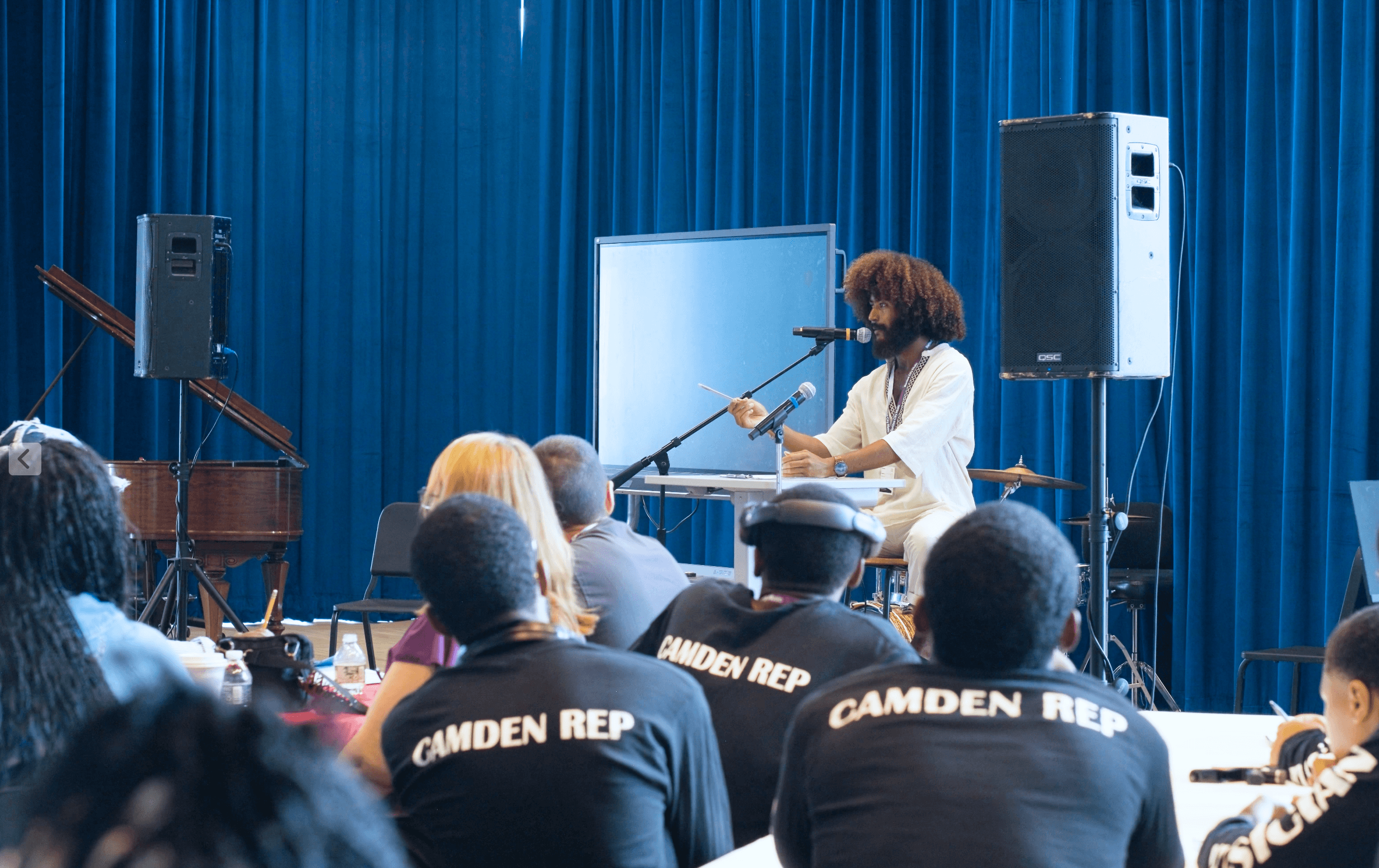 Performer playing drums on stage with blue curtains, audience watching, some wearing Camden Rep shirts.