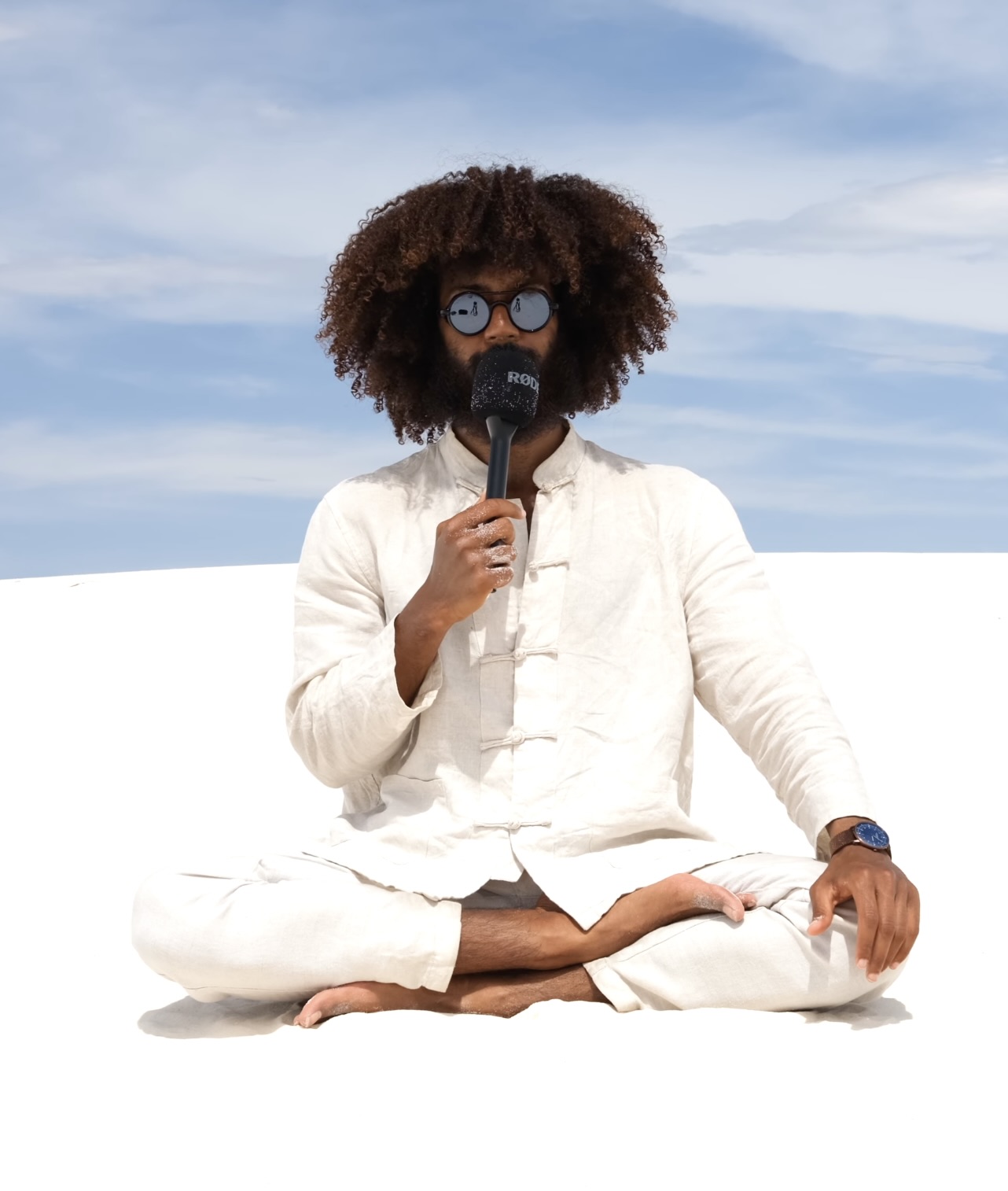 Person with curly hair, glasses, and a beard, sitting cross-legged outdoors, holding a microphone, against a blue sky background.