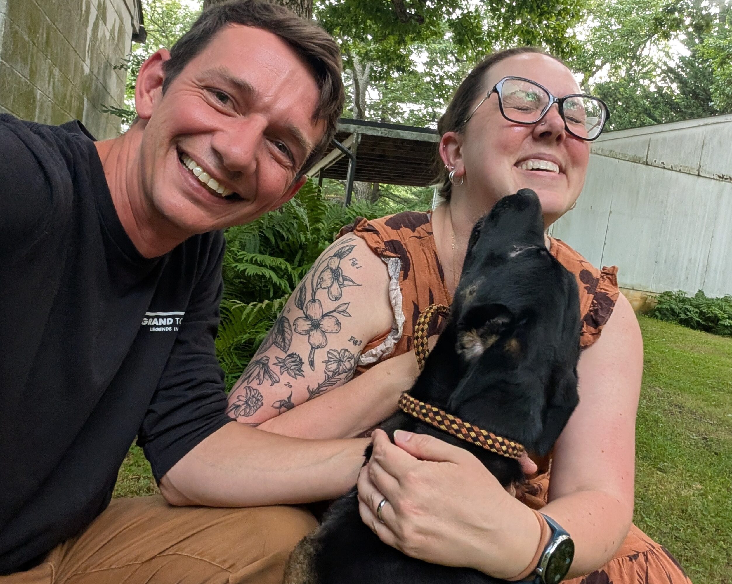 Two people smiling outdoors with a black dog, one person wearing glasses and a tattooed arm, greenery in background.