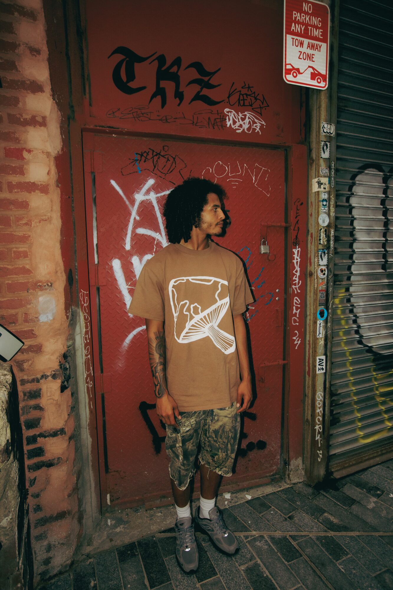 Young man with curly hair stands in front of a red graffiti-covered door, wearing a brown t-shirt and patterned shorts.