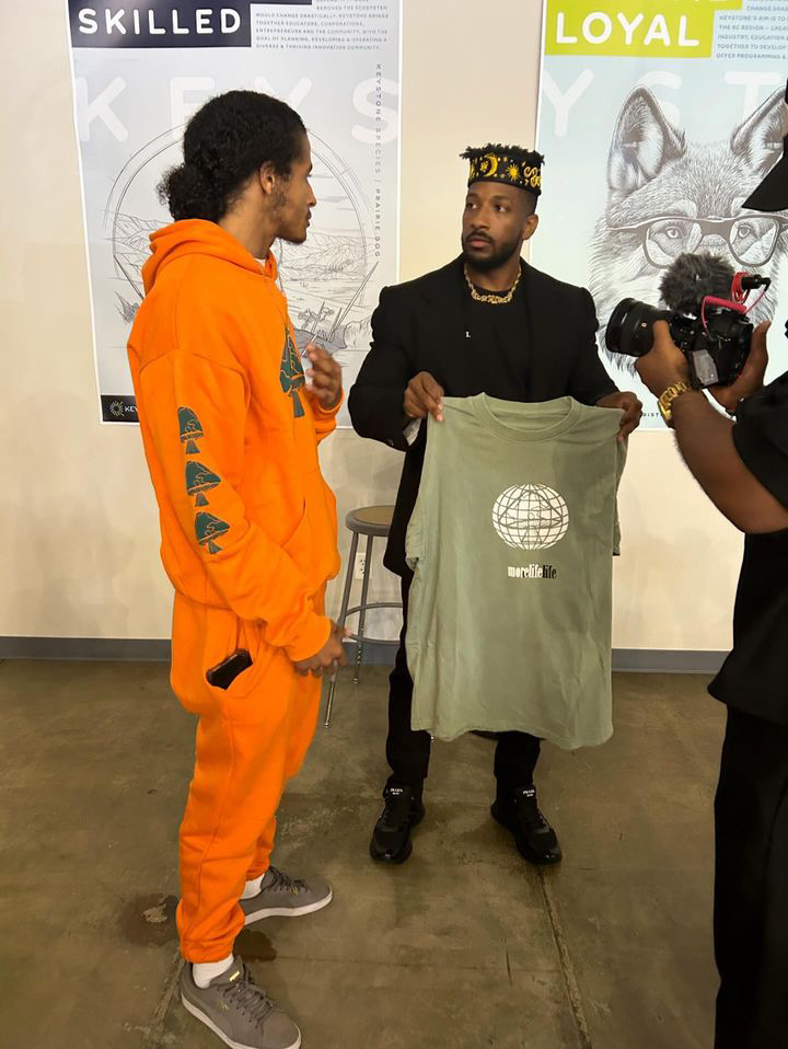Two men are engaged in conversation indoors, one holding a t-shirt with a globe logo, while a camera operator films them.