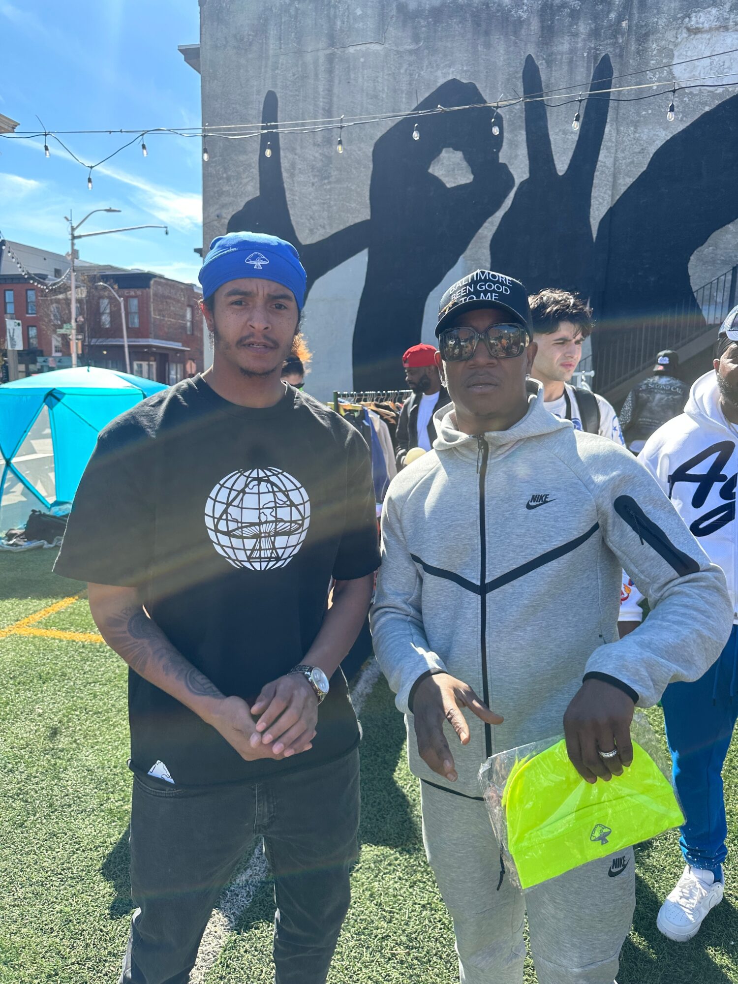 Two young men standing outdoors in front of a large mural with hand gestures, one wearing a black t-shirt and the other a gray jacket, holding a yellow bag.