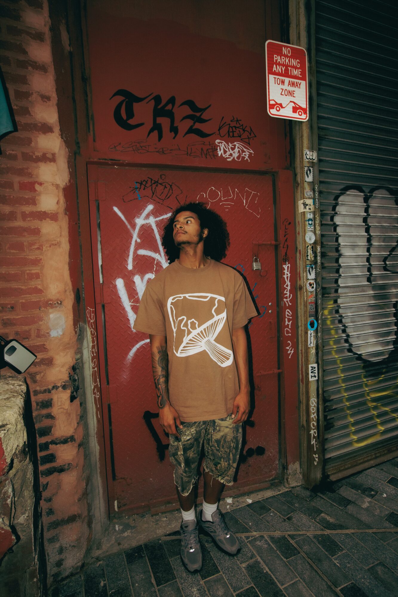 Young person with curly hair stands against a graffiti-covered red door in an urban setting, holding a phone in one hand.