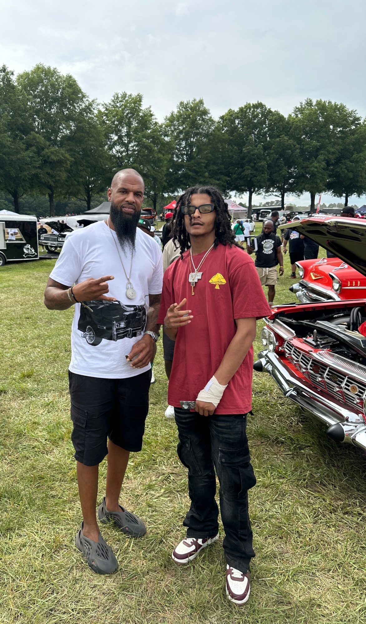 Two men standing outdoors near classic cars, one wearing a white t-shirt and shorts, the other in a red t-shirt and jeans, both making hand gestures.