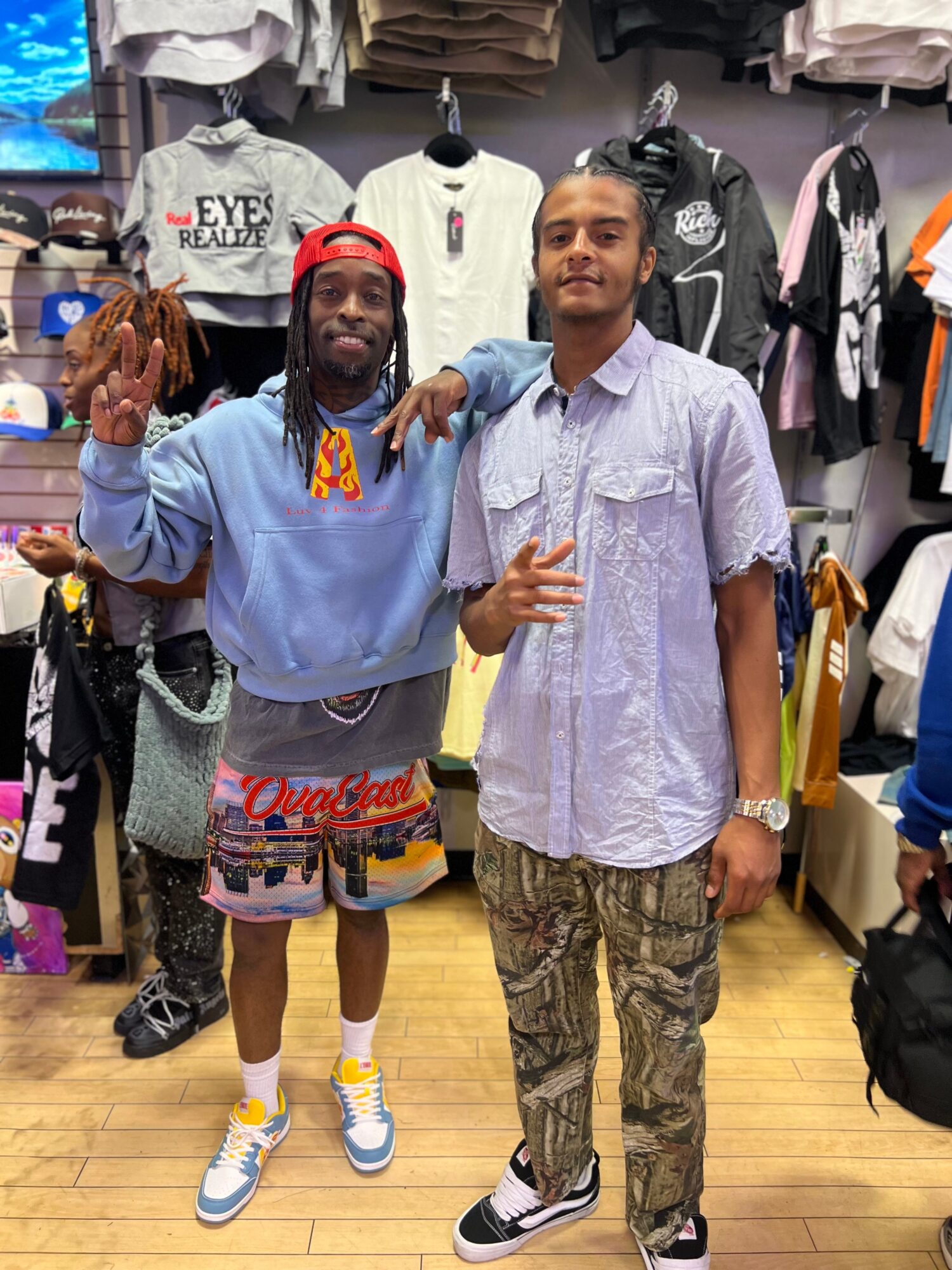Two young men stand in a clothing store, one with a red cap and colorful shorts, the other in a light purple shirt and patterned pants.